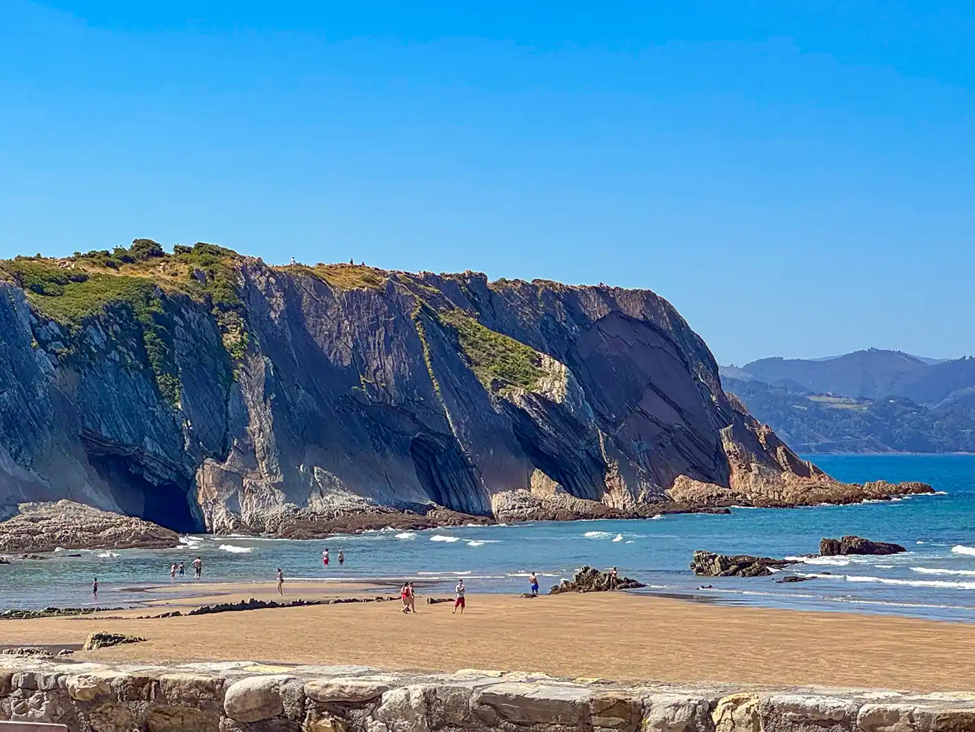 Menschen spazieren an einem Sandstrand mit felsigen Klippen und blauem Meer, im Hintergrund ist ein klarer Himmel zu sehen.
