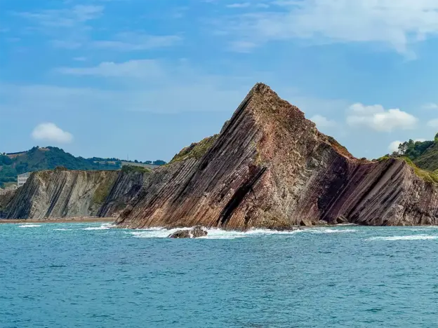 Unter einem teilweise bewölkten Himmel ragen zerklüftete, rötliche Felsformationen aus dem Meer empor.