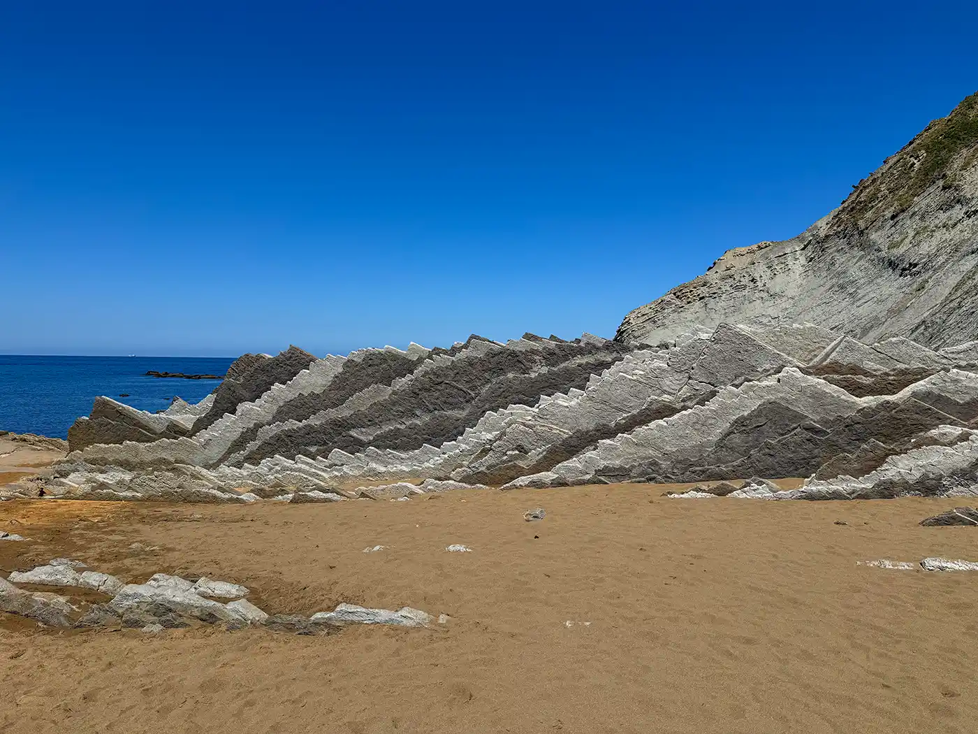 Unter einem strahlend blauen Himmel ragen neben dem Meer geschichtete Felsformationen aus einem Sandstrand empor.