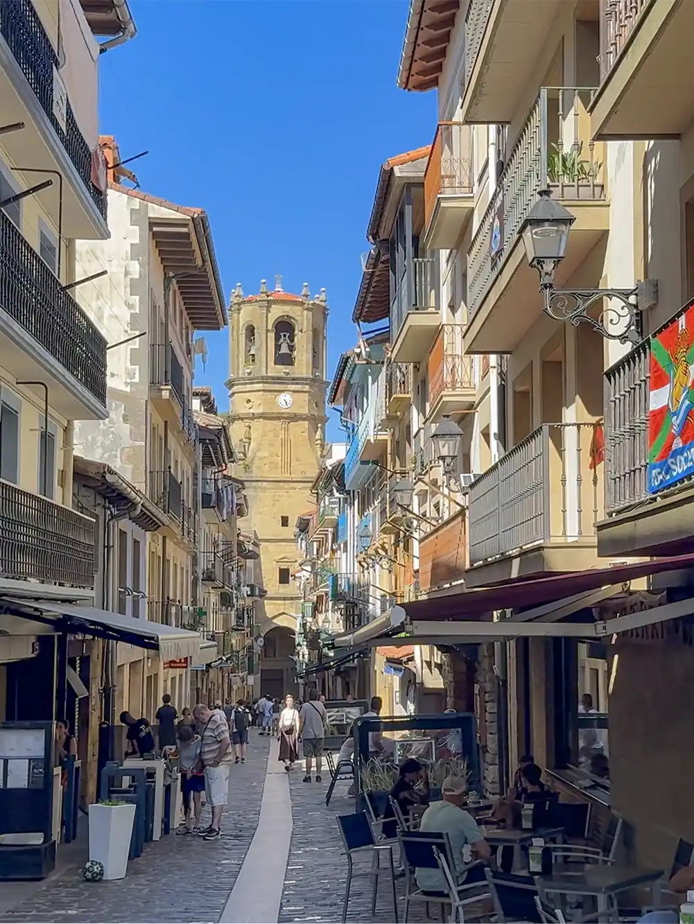 Getaria-Altstadt-Kale-Nagusia Eine schmale Straße mit Straßencafés, bunten Balkonen und einem Uhrturm unter strahlend blauem Himmel.