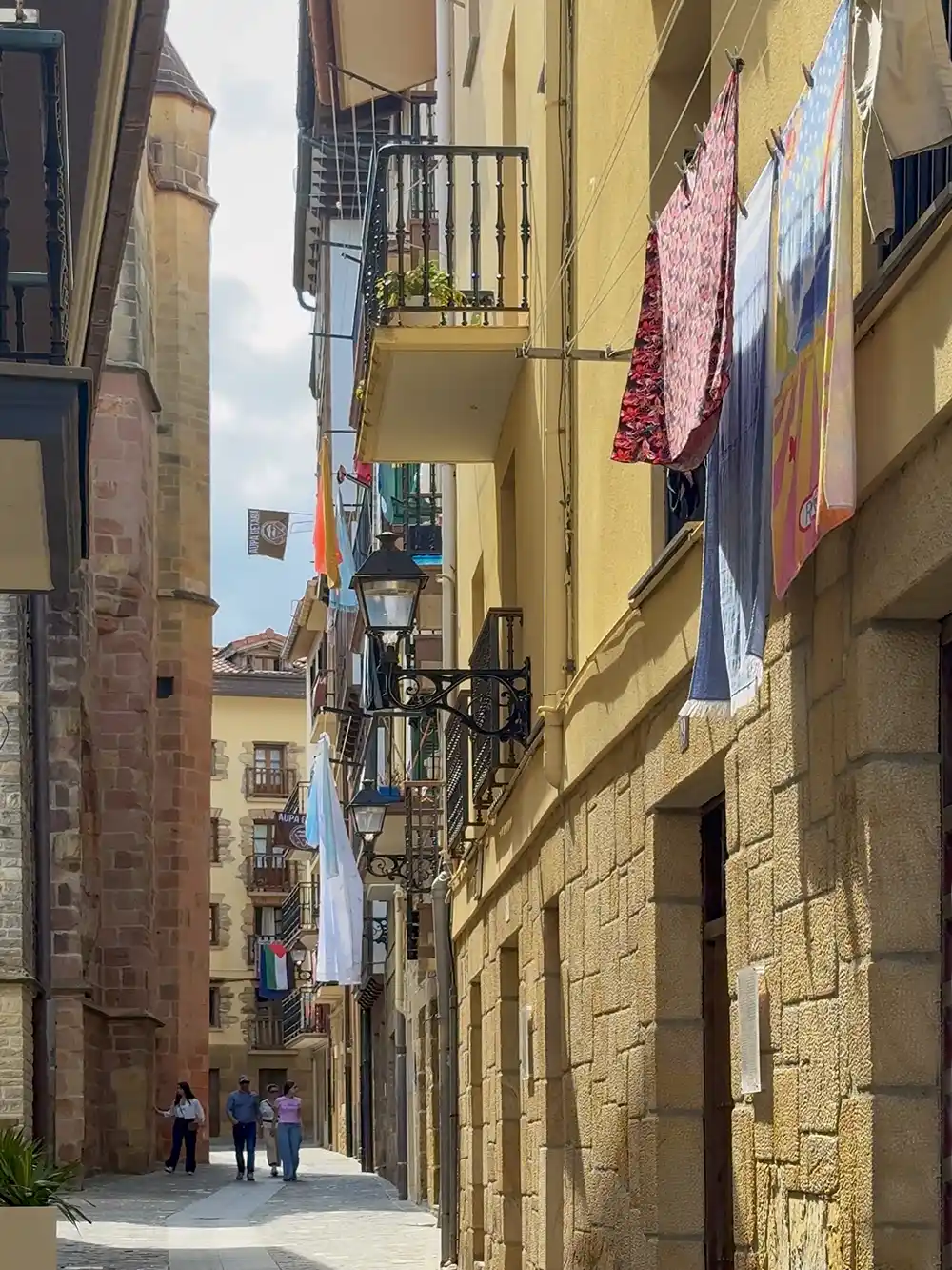 Getaria-Altstadt-Kirche-Waesche Eine schmale Straße mit aufgehängter Wäsche, Steinhäusern, Balkonen und drei Personen, die im Hintergrund spazieren gehen.