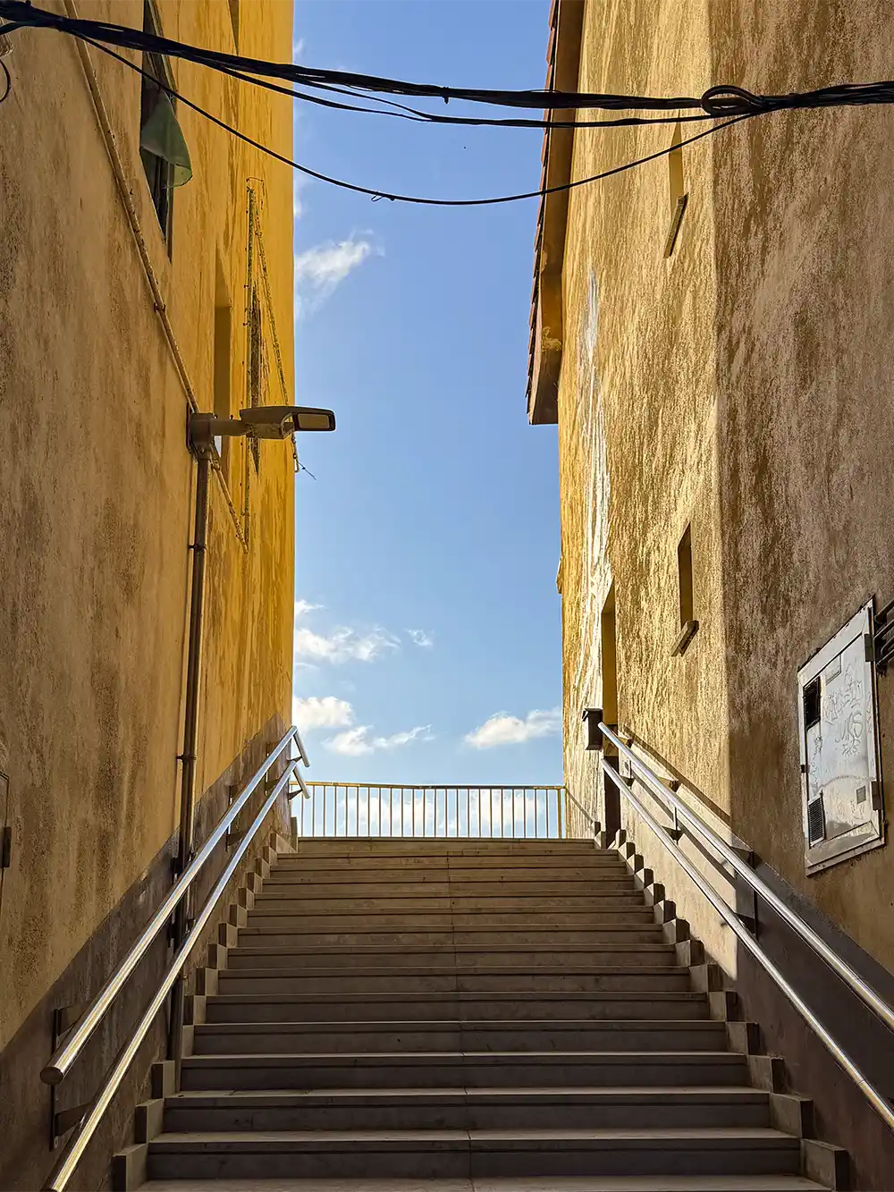 Getaria-Altstadt-Treppen Eine Steintreppe zwischen zwei gelben Gebäuden, die hinauf zum blauen Himmel mit Wolken und Oberleitungen führt.