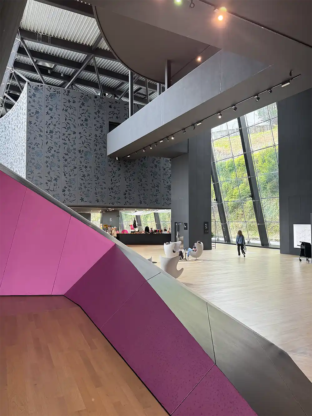 Getaria-Balenciaga-Museum-innen Moderne Museumslobby mit großen Fenstern, einer rosa Wand mit geometrischen Mustern und zwei Personen, die darin entlanggehen.