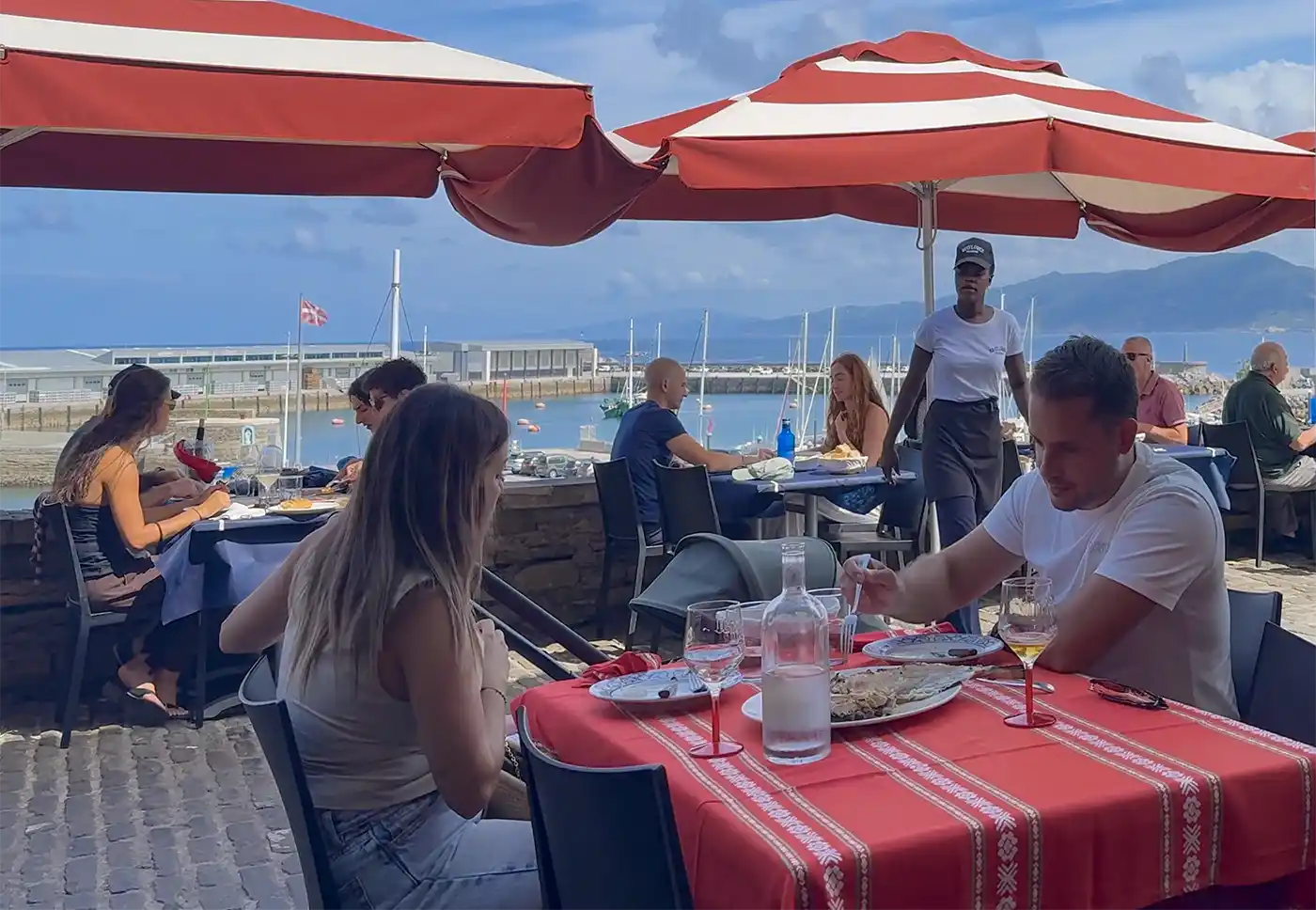Getaria-Baskenland-Fisch-Restaurant-Mayflower Menschen, die im Freien unter roten Sonnenschirmen an einem Yachthafen speisen, im Hintergrund Boote und Berge.