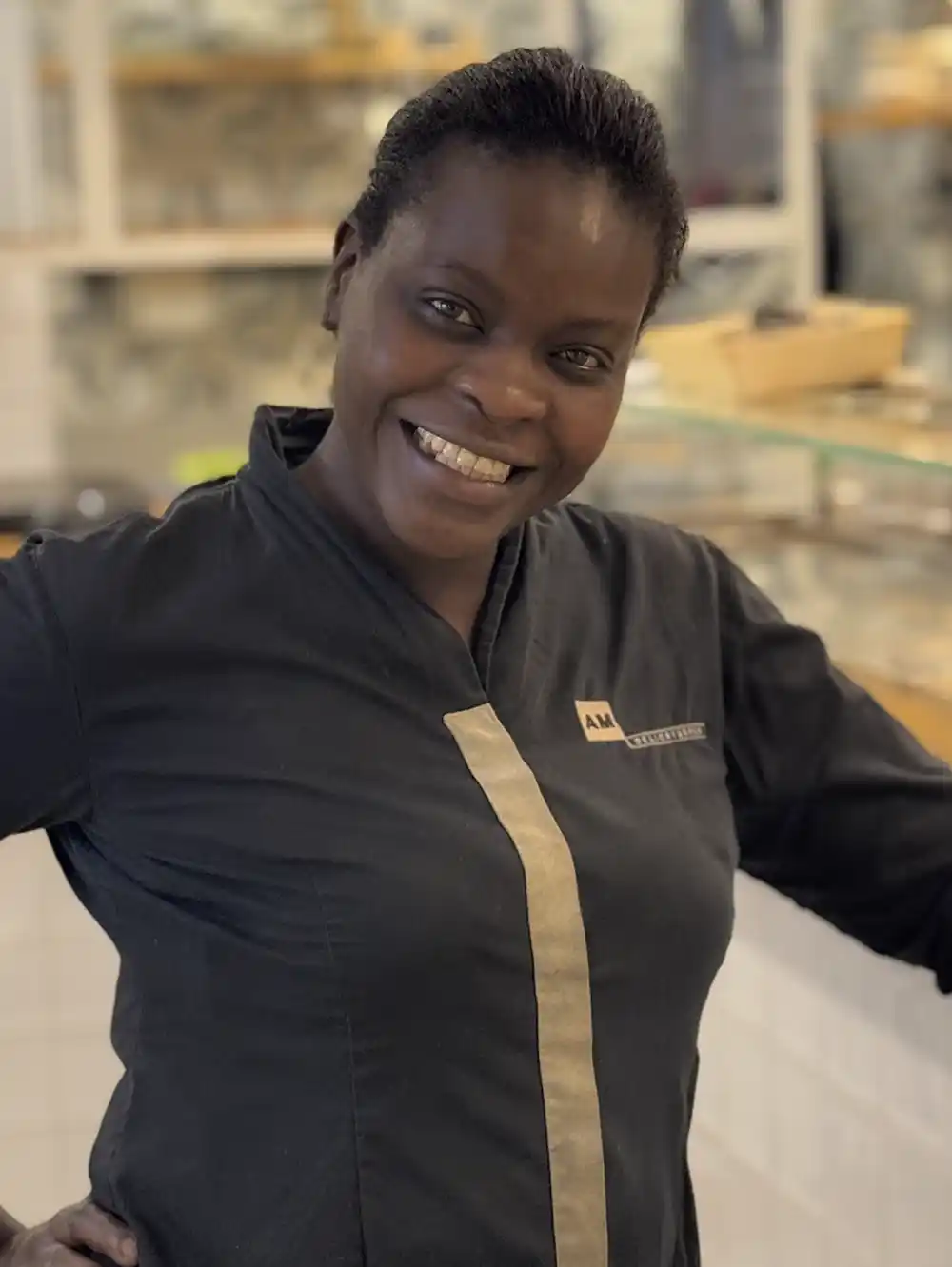 Getaria-Baskenland-Koechin-Salla-AM-Delicatessen Eine Frau in einer dunklen Kochjacke lächelt in einer Bäckerei, im Hintergrund sind Regale und Backwaren zu sehen.