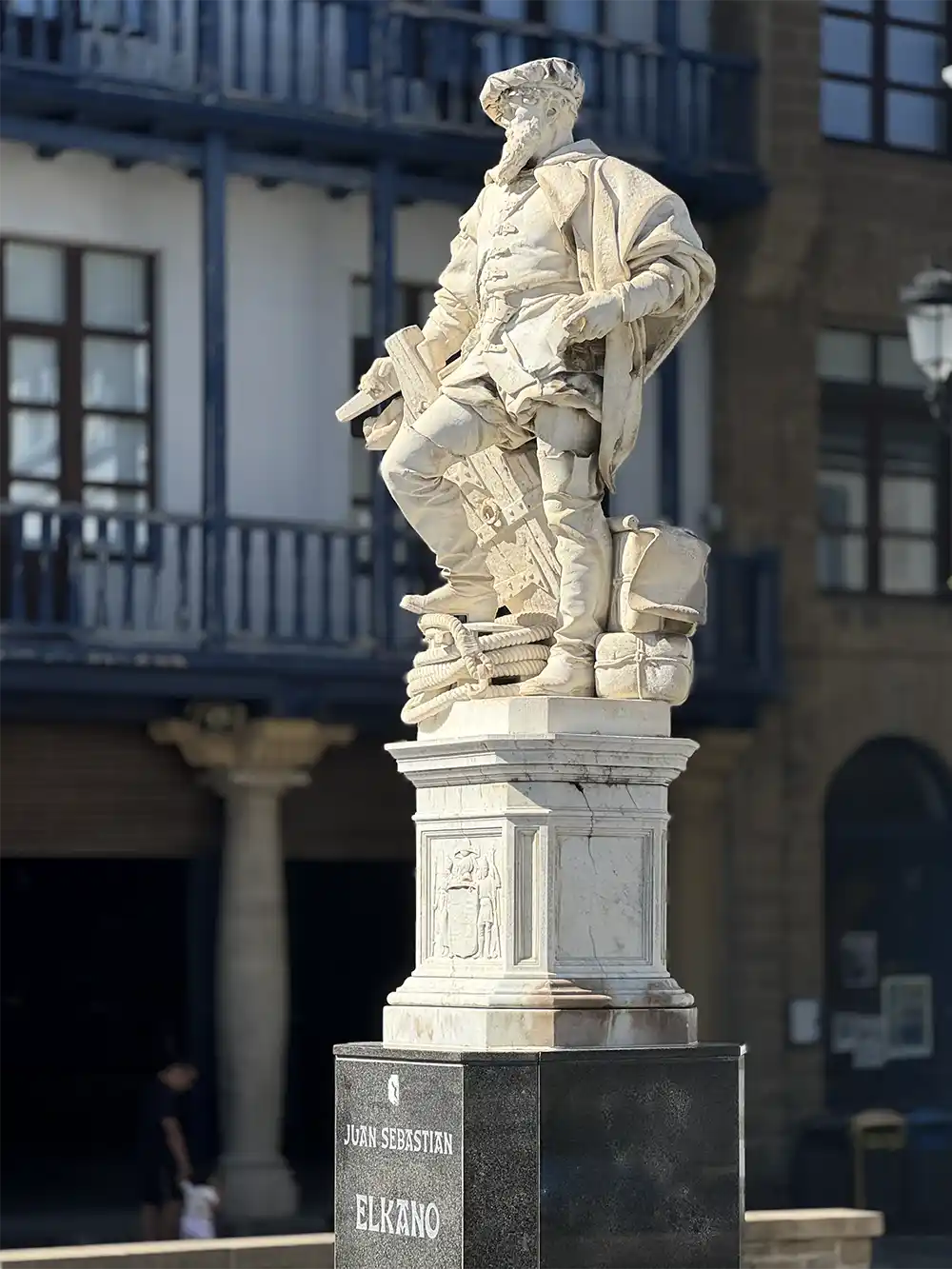 Getaria-Elkano-Denkmal-Marmor Statue eines Mannes in historischer Kleidung, der auf einem Sockel mit der Aufschrift „JUAN SEBASTIAN ELKANO“ im Freien steht.
