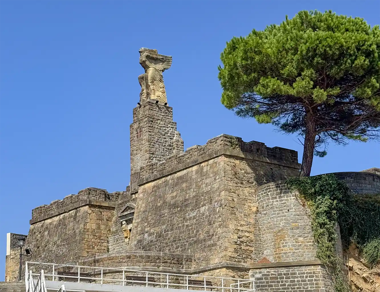 Getaria-Elkano-Denkmal Eine alte Steinfestung mit einer Engels-Statue auf der Spitze, neben einem großen Baum unter strahlend blauem Himmel.