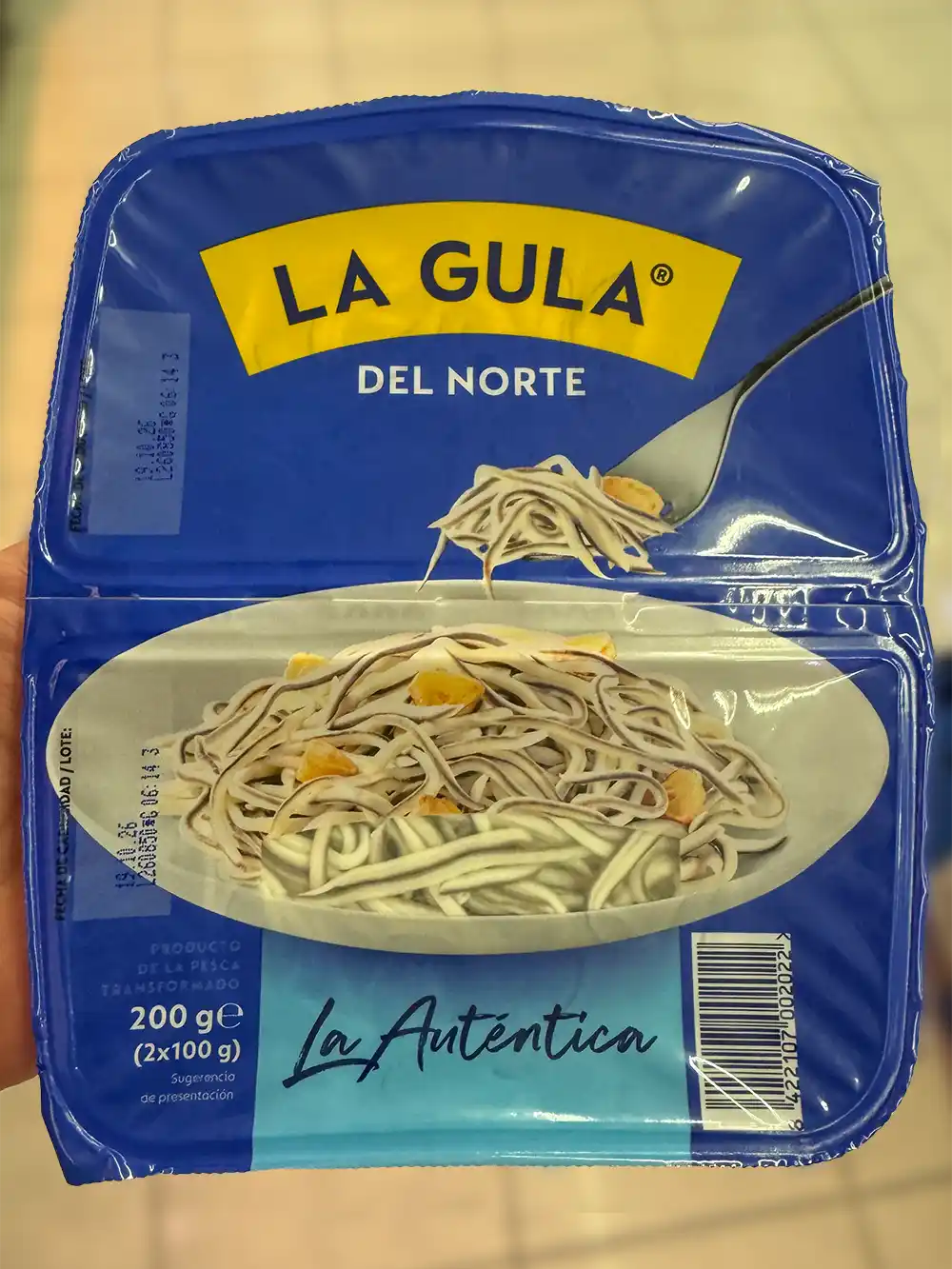 Verpackung von „La Gula del Norte“ mit der Abbildung von künstlichen Baby-Aalen und Knoblauch auf einem Teller, der in einer Hand gehalten wird.