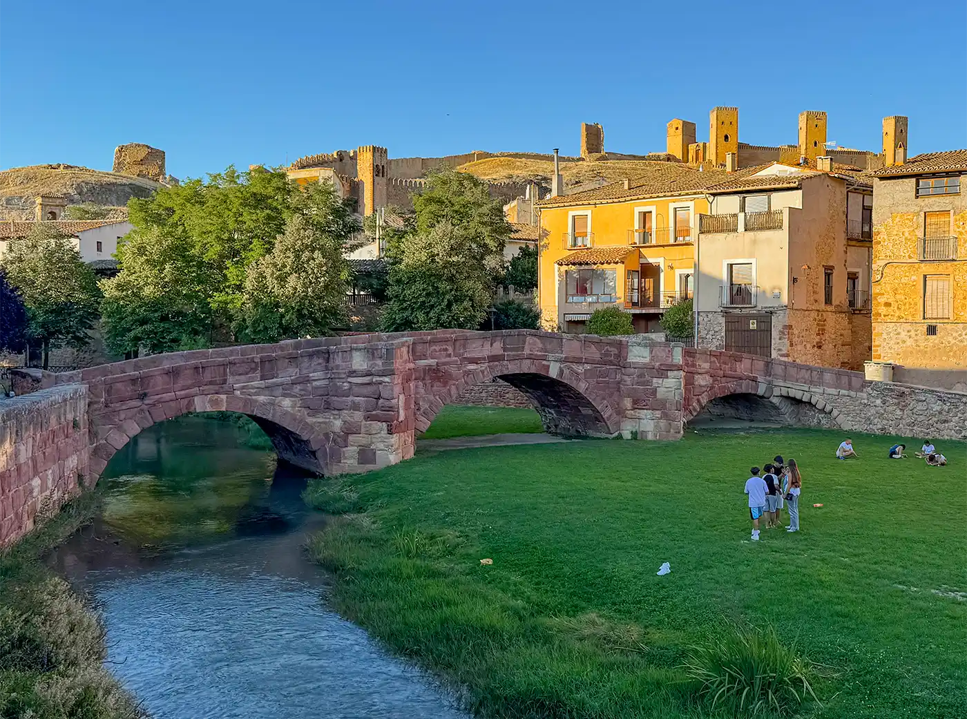 Sandsteinfarbene Mittelalterliche Brücke mit 3 Bögen, links fließt ein Bach durch, unter den beiden anderen ist Rasen, auf dem Kinder spielen