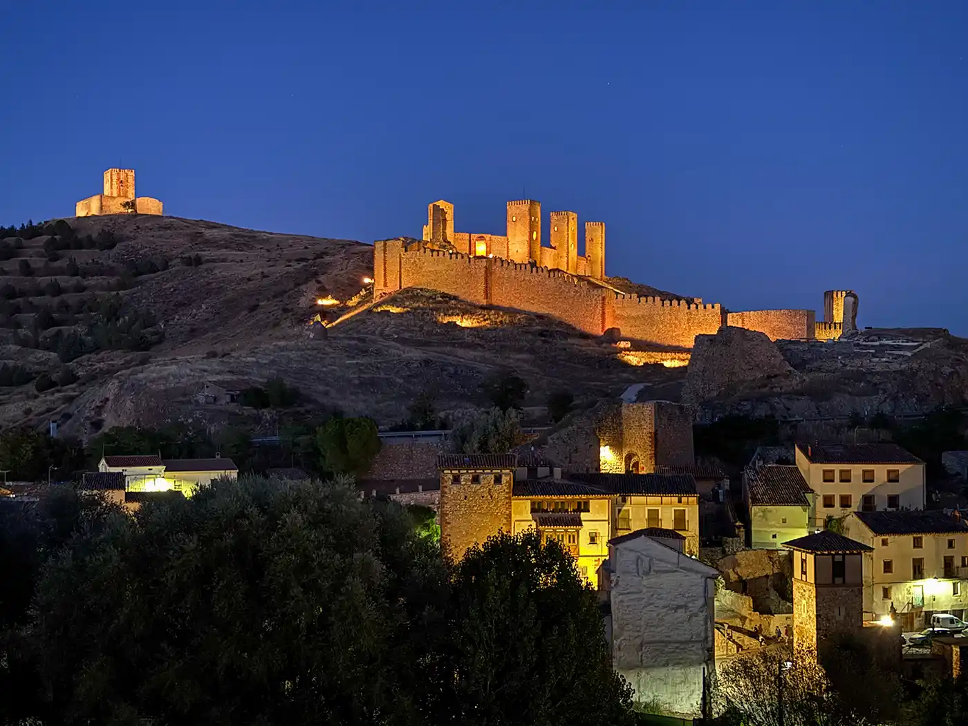 Die mittelalterliche Burg Molina de Aragon auf einem Hügel, die nachts beleuchtet ist und einen kleinen Ort mit Steinhäusern überragt.