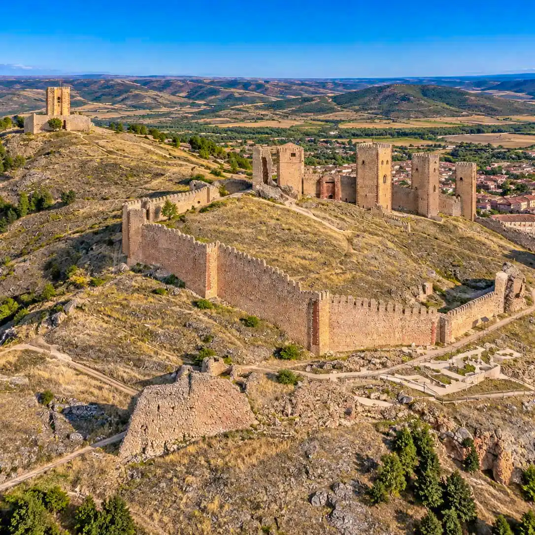 Ruinen einer mittelalterlichen Burg auf einem Hügel mit Steinmauern, Türmen und einer weitläufigen grünen Landschaft im Hintergrund unter einem strahlend blauen Himmel.