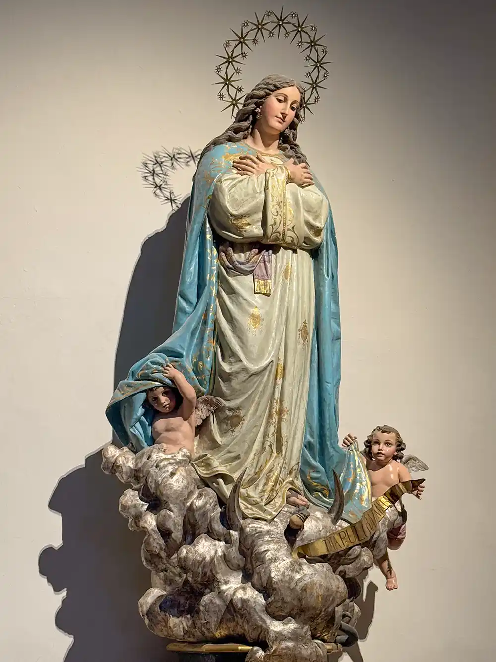 Statue der Maria mit blauem Gewand, gefalteten Händen, Heiligenschein und zwei Cherubim auf Wolken zu ihren Füßen.