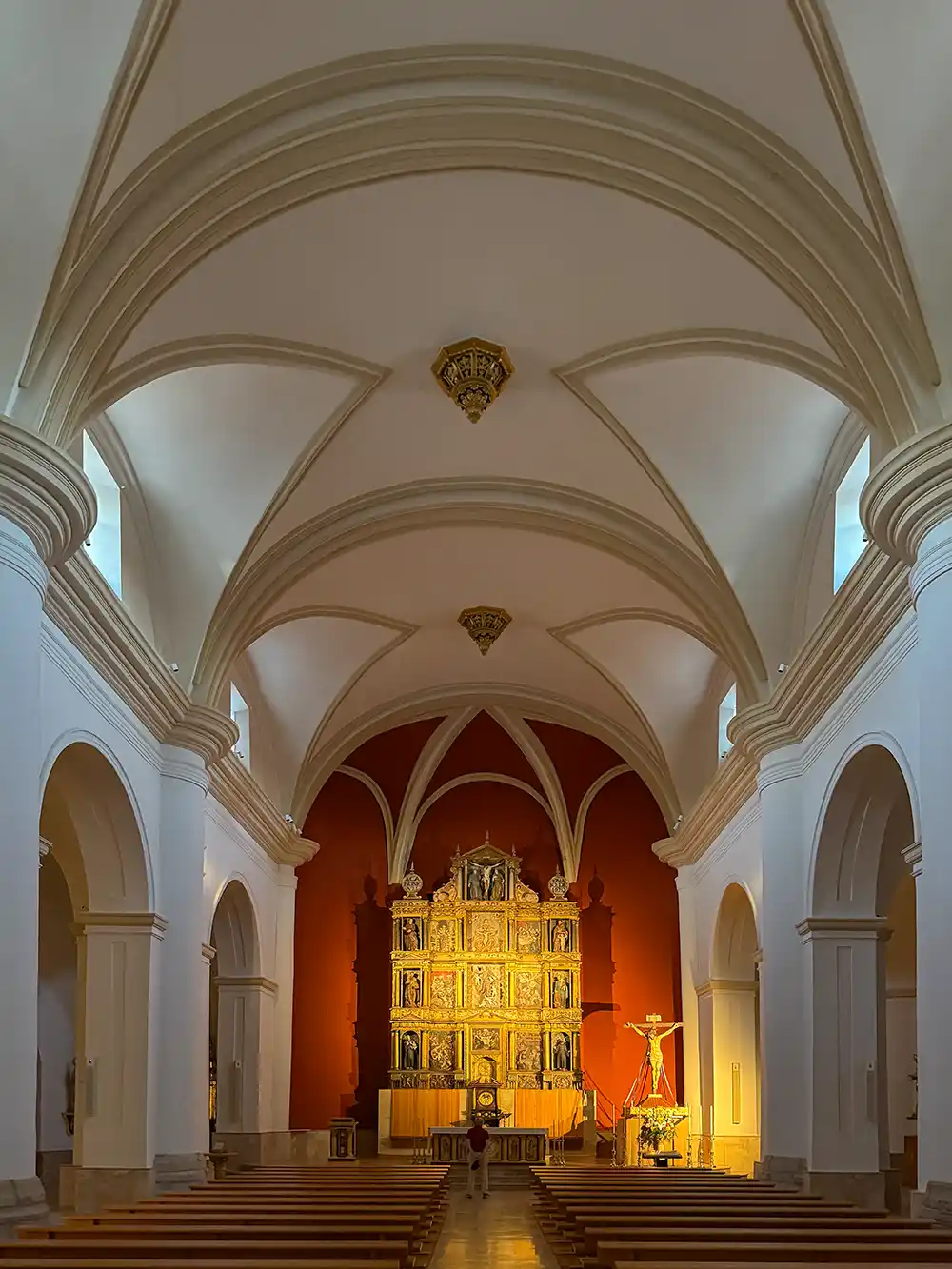 Ein Kircheninneres mit Gewölbedecken, Holzbänken und einem reich verzierten Altar vor einer roten Wand.