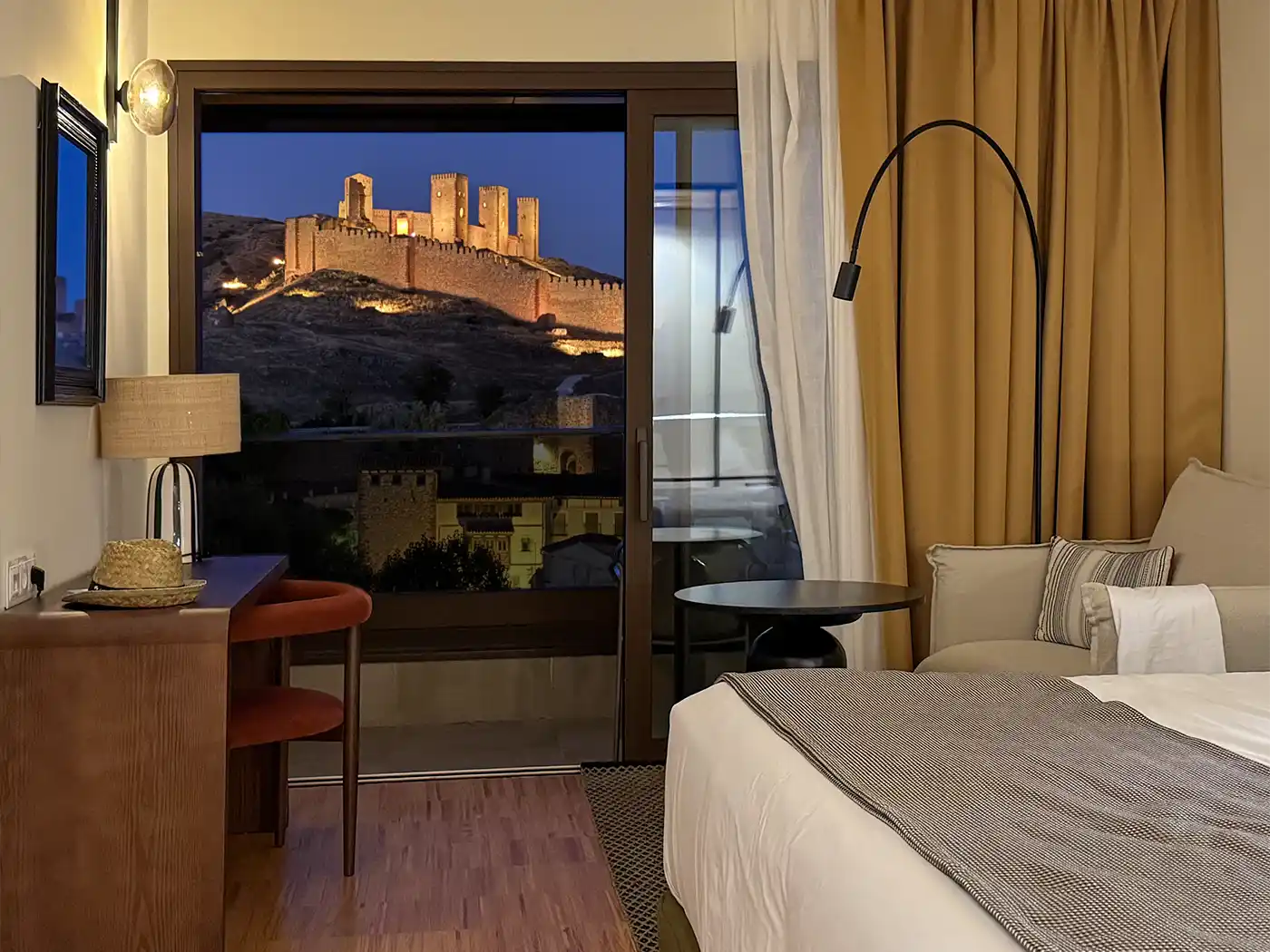 Hotelzimmer mit Bett, Schreibtisch und Blick aus dem Fenster auf eine beleuchtete Burg auf einem Hügel in der Abenddämmerung.