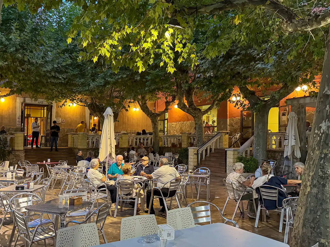 Am Abend sitzen die Menschen an den Tischen der Straßencafés unter grünen Bäumen und unterhalten sich, während warmes Licht leuchtet.