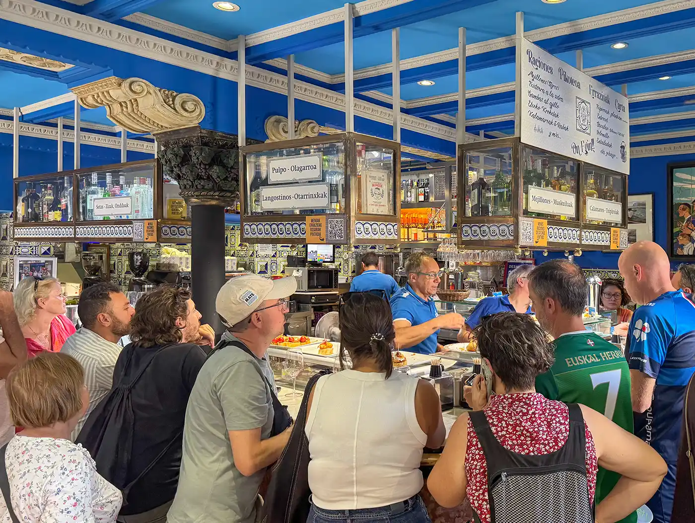 Pintxo-Bar-Bilbao Eine Gruppe von Menschen versammelt sich um eine belebte Marktbar mit blauen Decken und handgeschriebenen Speisekarten.