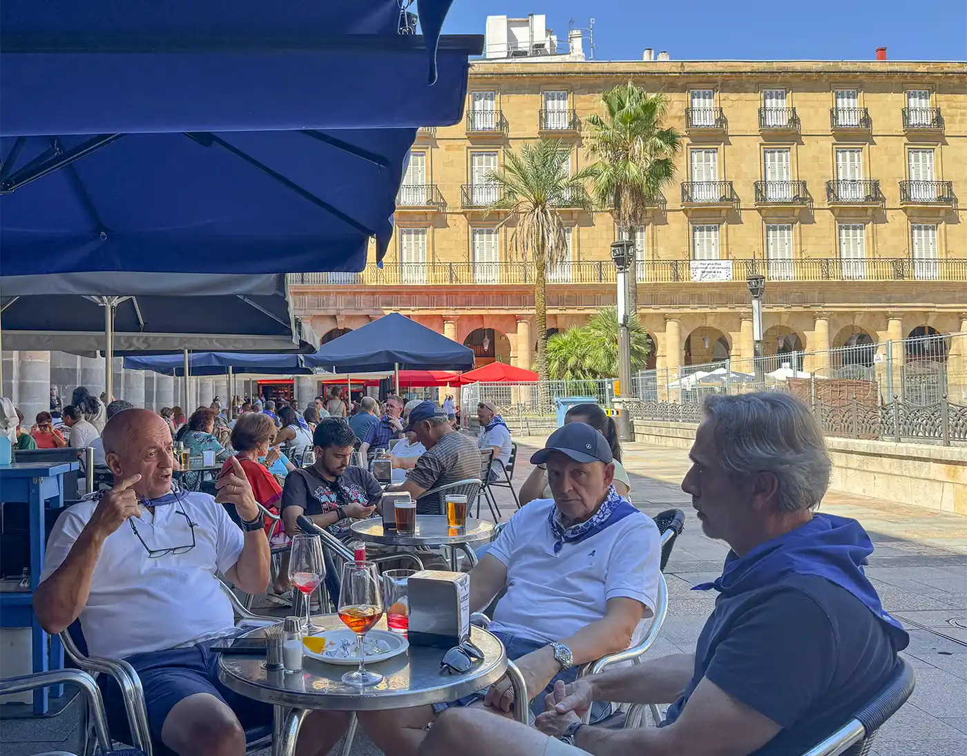Pintxo-Cafe-Bar-Bilbao-Plaza-Nueva Drei Männer sitzen an einem Tisch im Straßencafé, vor dem Hintergrund von Getränken, anderen Menschen und historischen Gebäuden.