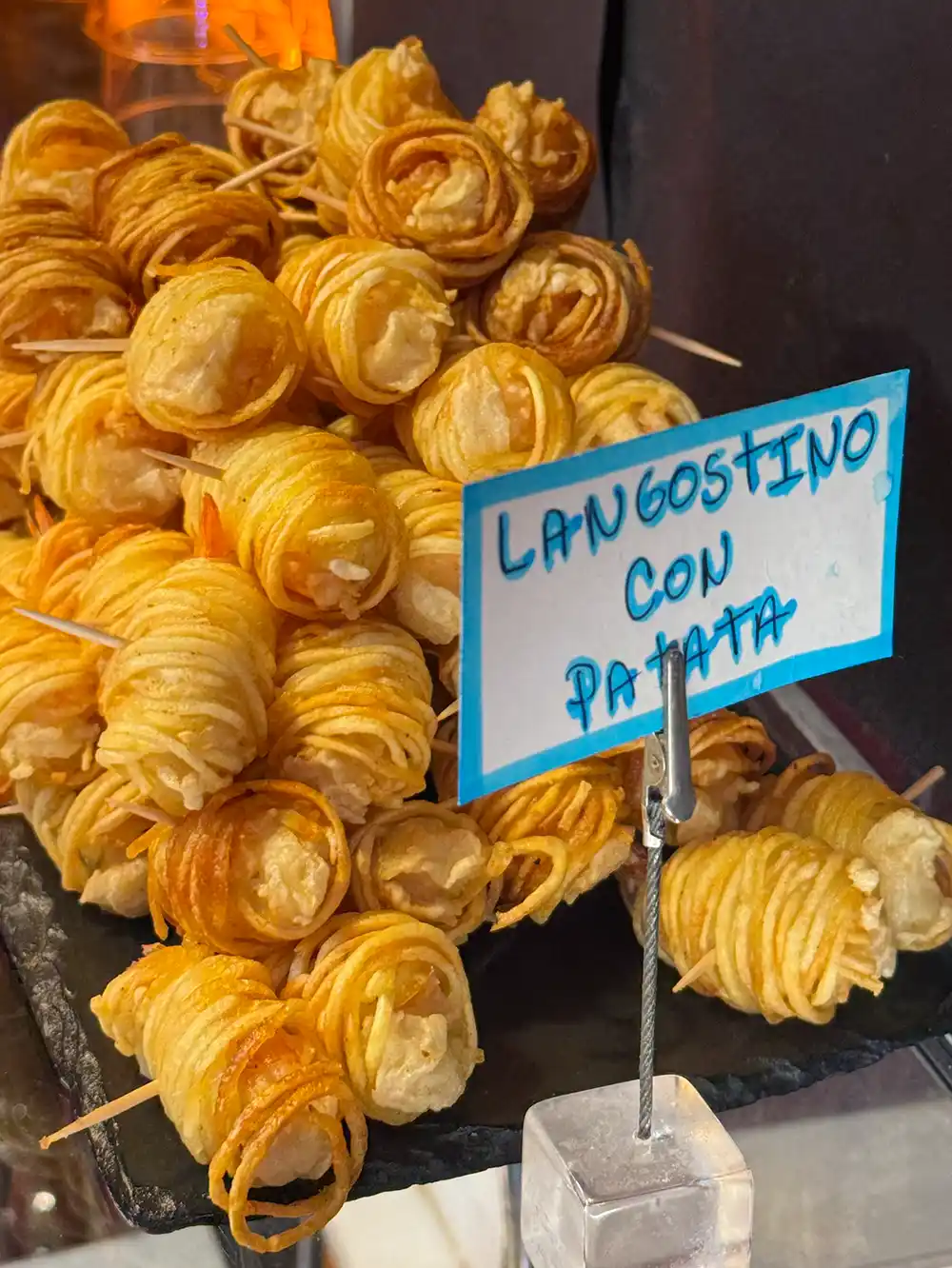 Gebratene Garnelen, eingewickelt in Kartoffelstreifen, auf einem Tablett, mit einem handgeschriebenen Schild mit der Aufschrift „Langostino con patata“.