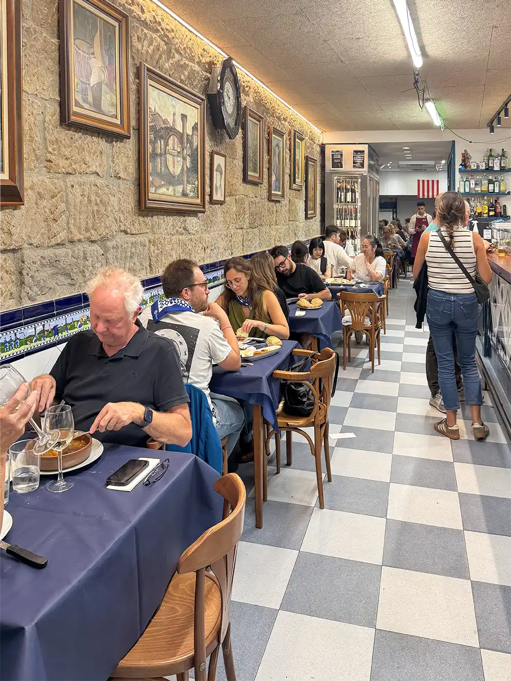 Gäste, die in einem gemütlichen Restaurant mit blauen Tischdecken, Kunstwerken an den Steinwänden und einem langen, gefliesten Gang speisen.