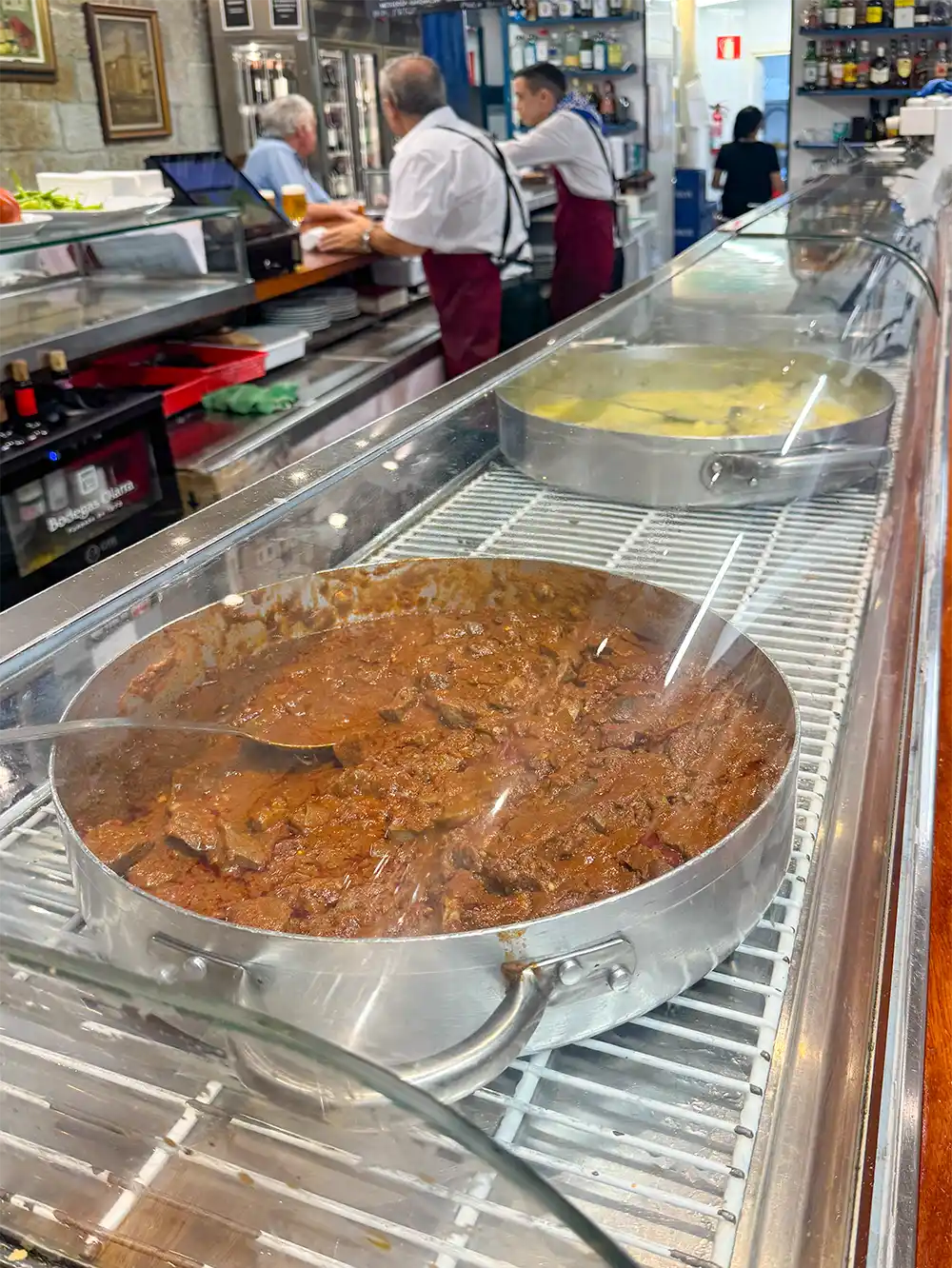 Zwei große Pfannen mit Essen, eine mit Eintopf und eine mit Suppe, stehen hinter einer Glasvitrine in einem Feinkostladen ausgestellt.