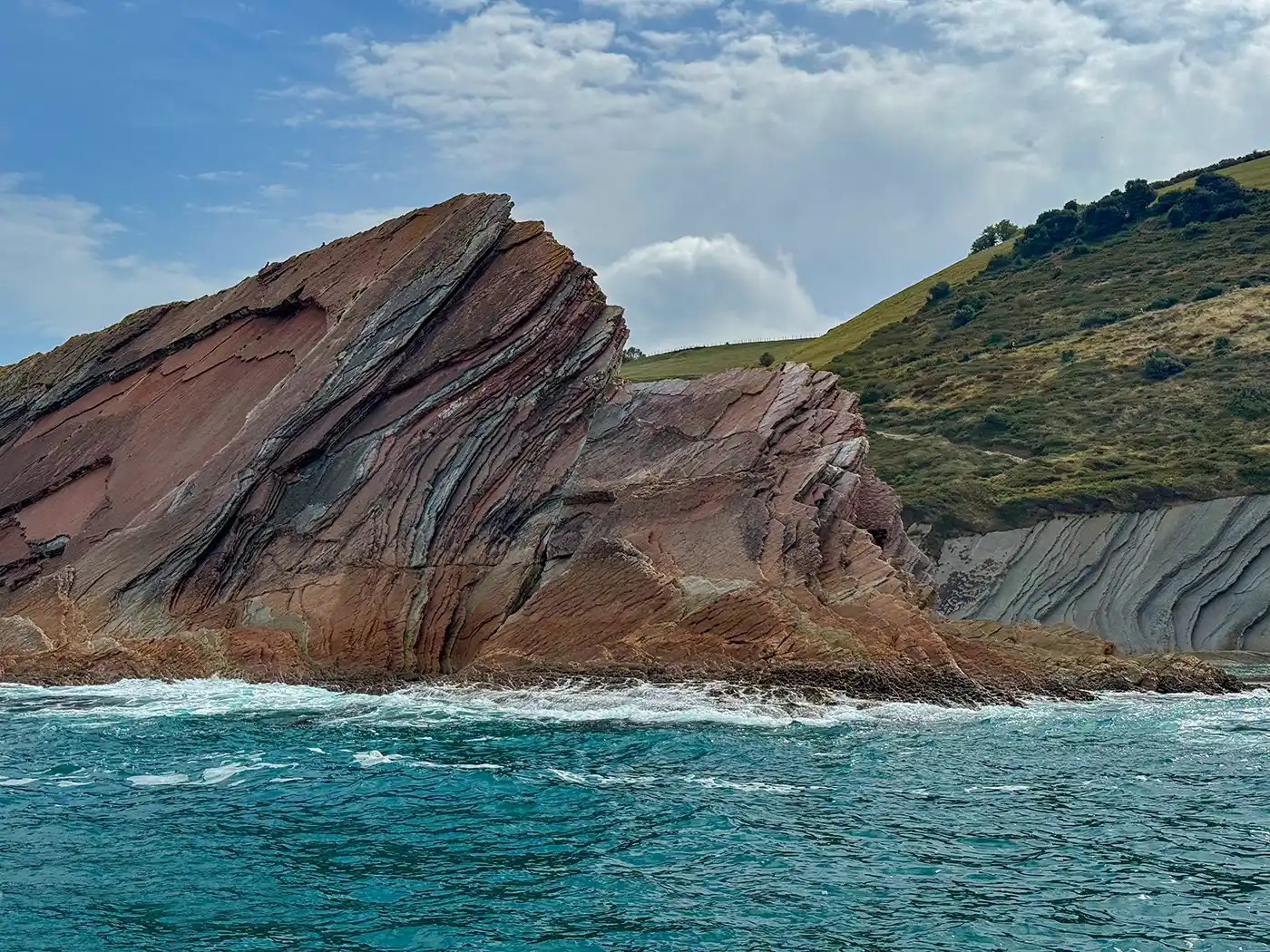 Eine schräg stehende Felsformation am Meer, brechende Wellen, ein grasbewachsener Hügel und ein bewölkter Himmel im Hintergrund.