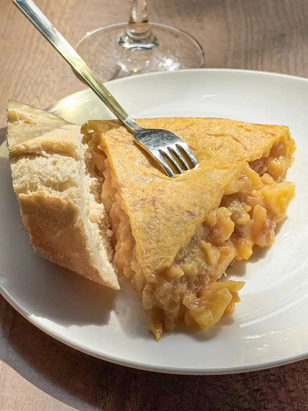 Ein Stück spanisches Omelett mit Kartoffeln auf einem weißen Teller, serviert mit einem Stück Brot und einer Gabel darauf.