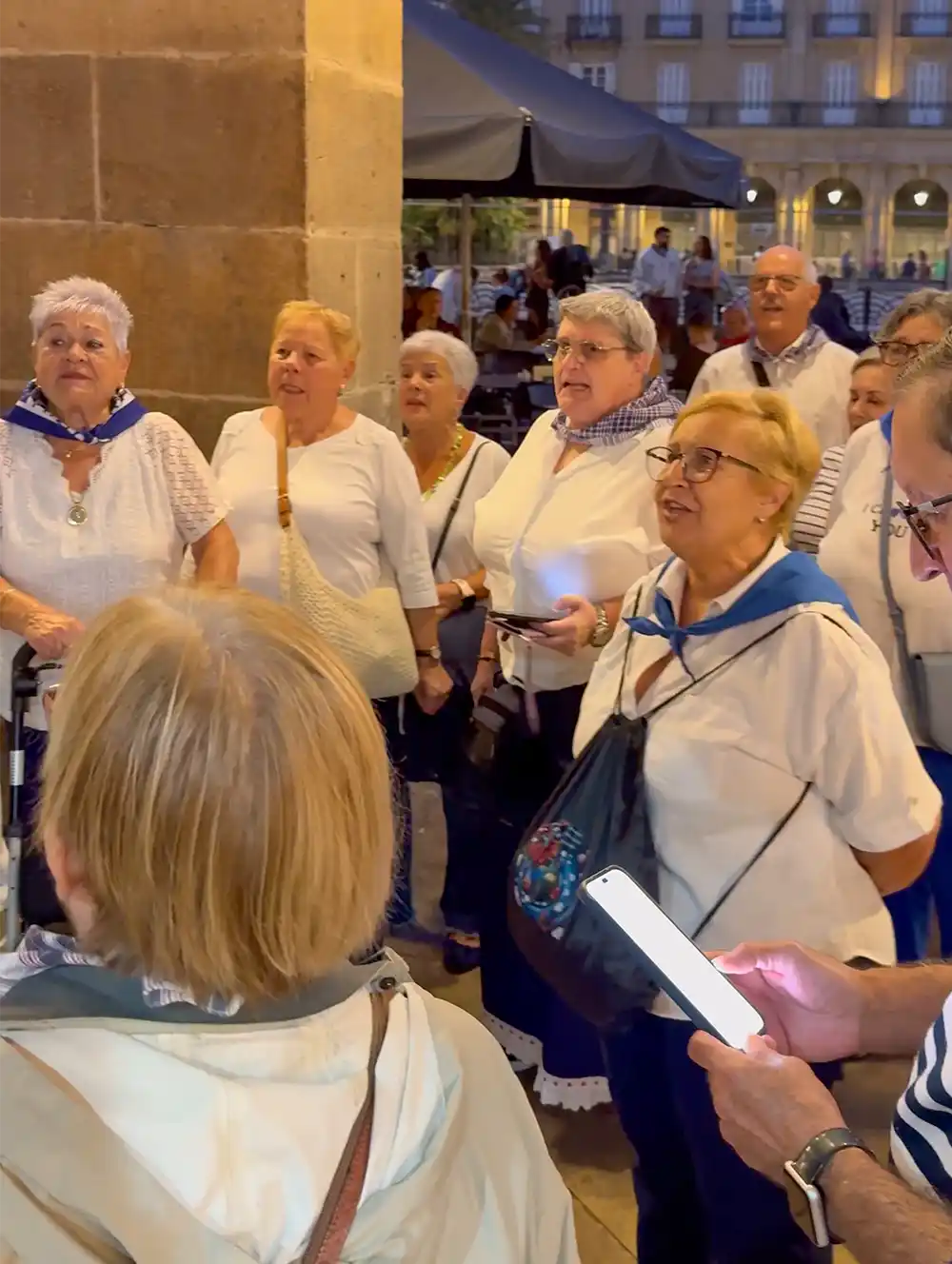 Eine Gruppe älterer Menschen steht gemeinsam im Freien, einige singen, viele tragen weiße Kleidung und blaue Schals.