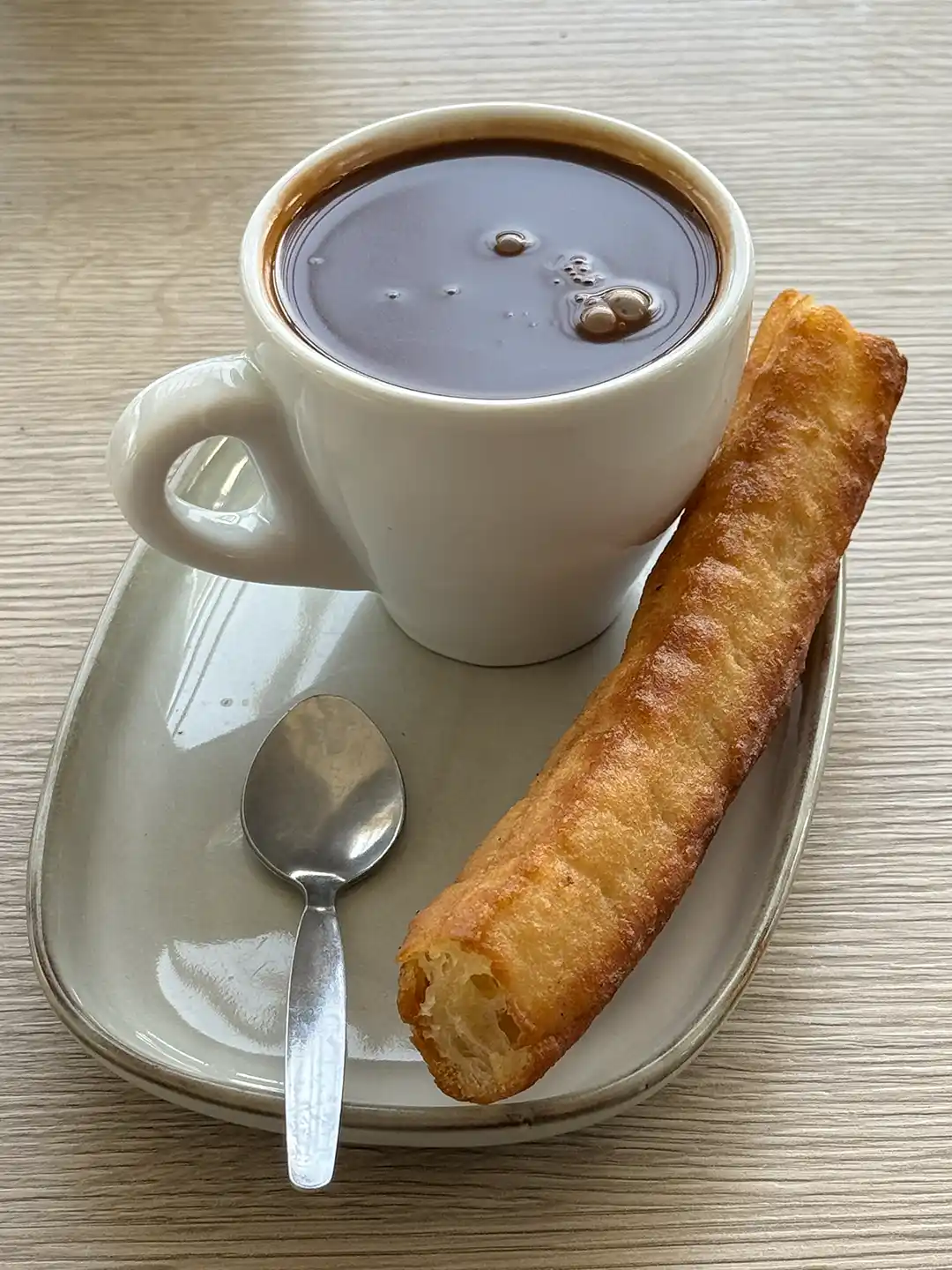 Eine Tasse heiße Schokolade mit einem Churro und einem Löffel auf einem kleinen Teller.