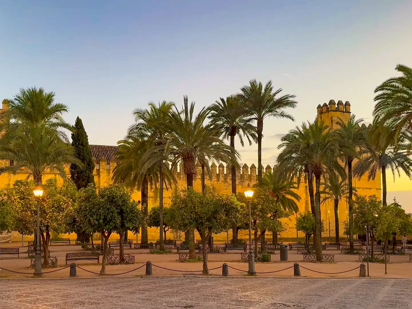 Cordoba-Alcazar-de-los-Reyes-Cristianos Palmen vor einer historischen Festungsmauer aus Stein bei Sonnenuntergang.