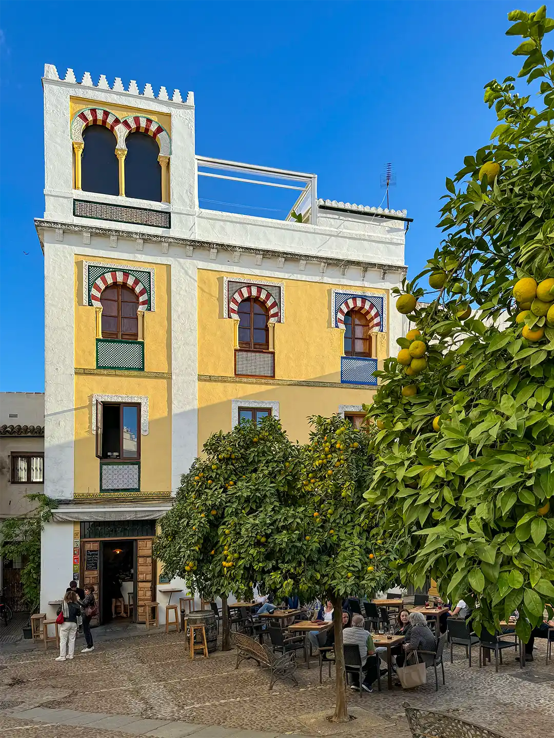 Cordoba-Altstadt Café im Freien in einem Innenhof mit einem hohen, gelben Gebäude, das sich durch Bögen im maurischen Stil und Orangenbäume auszeichnet.