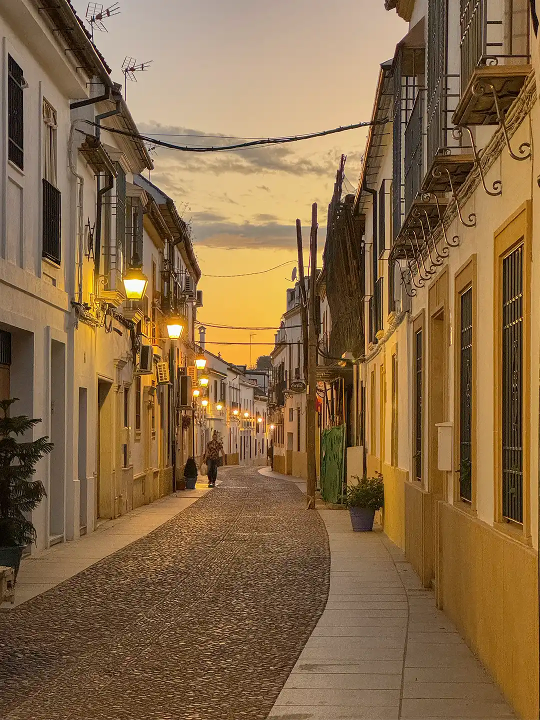 Cordoba-Juderia-Gasse Eine schmale Kopfsteinpflasterstraße, gesäumt von weißen Gebäuden, die bei Sonnenuntergang im warmen Licht der Straßenlaternen erstrahlen.