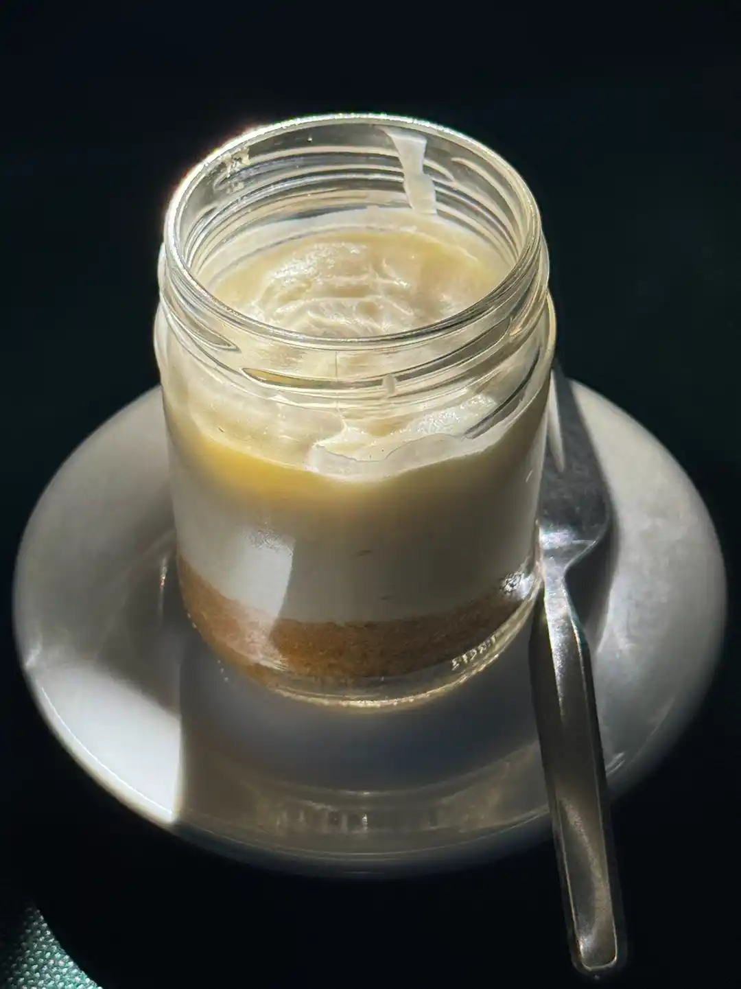 Ein geschichtetes Dessert in einem kleinen Glas auf einer weißen Untertasse, dazu ein Löffel.