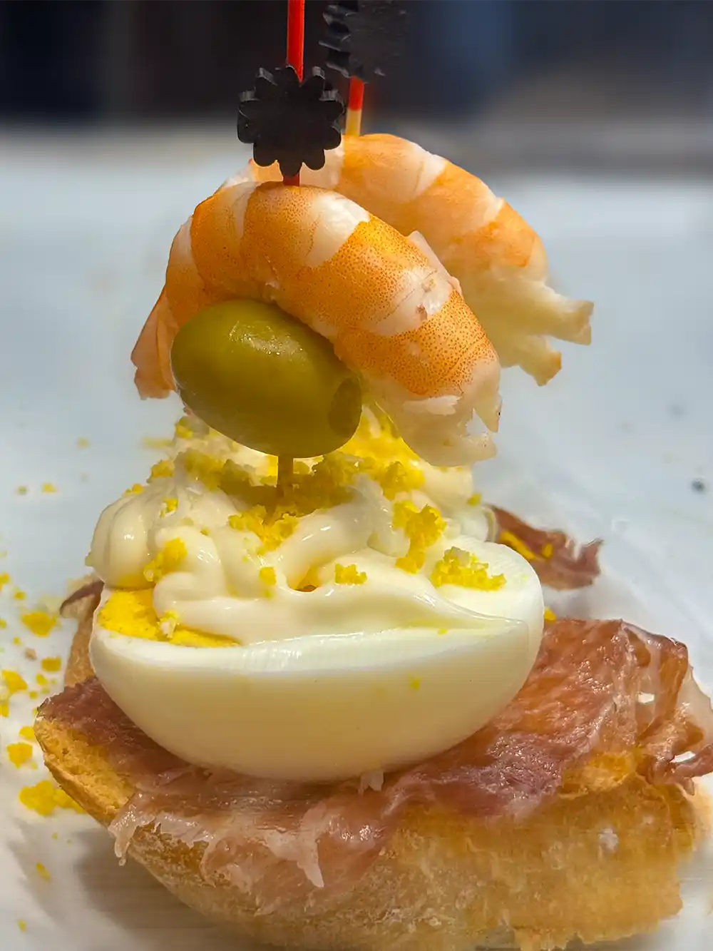 Ein halbgekochtes Ei auf Schinken und Brot, garniert mit Garnelen, einer Olive, Mayonnaise und geriebenem Eigelb.