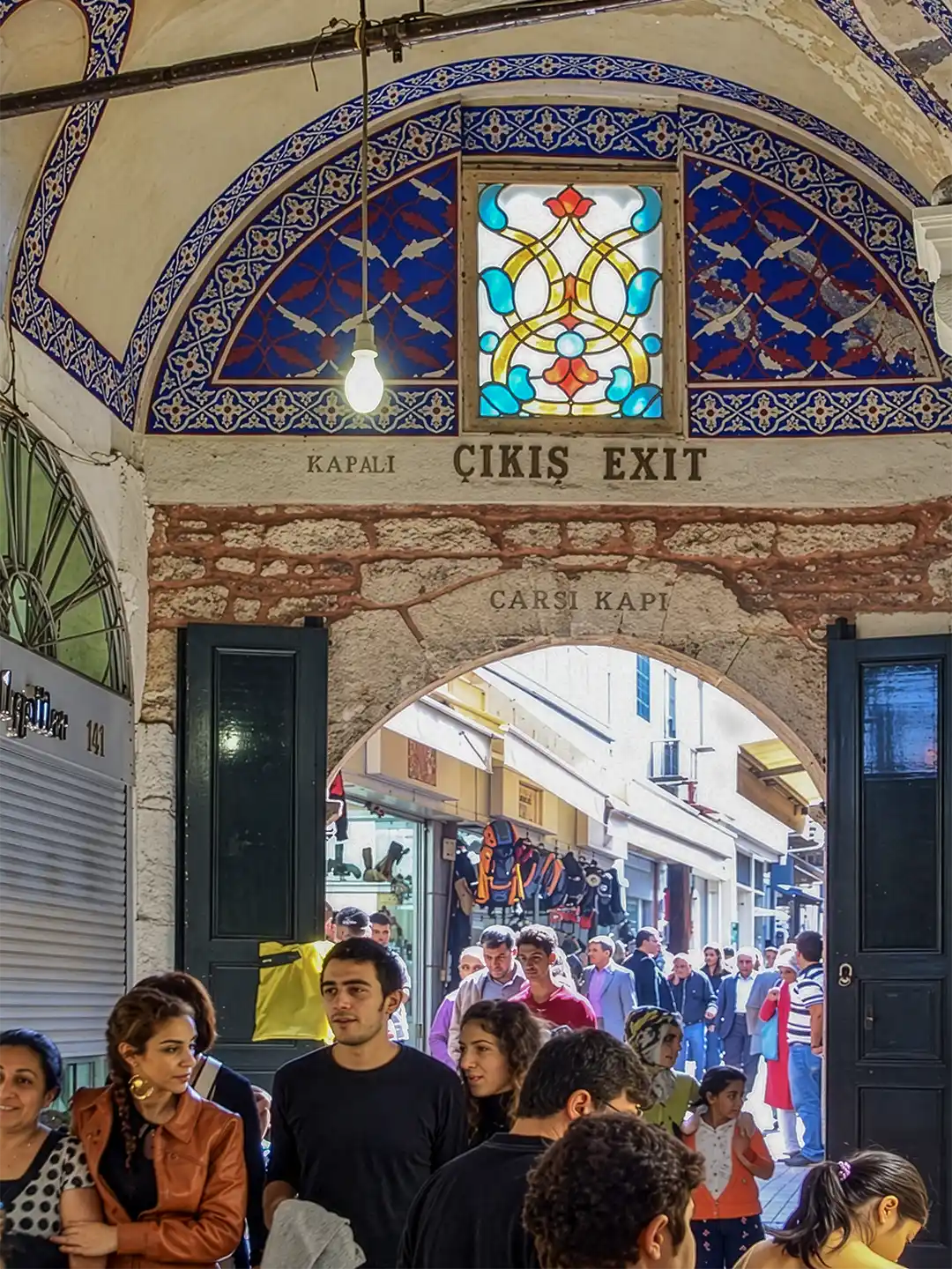 Istanbul-Basar-Ausgang Eine Menschenmenge geht durch ein gewölbtes Steintor mit Buntglasfenstern und einem Schild mit der Aufschrift KAPALI ÇIKIŞ EXIT.