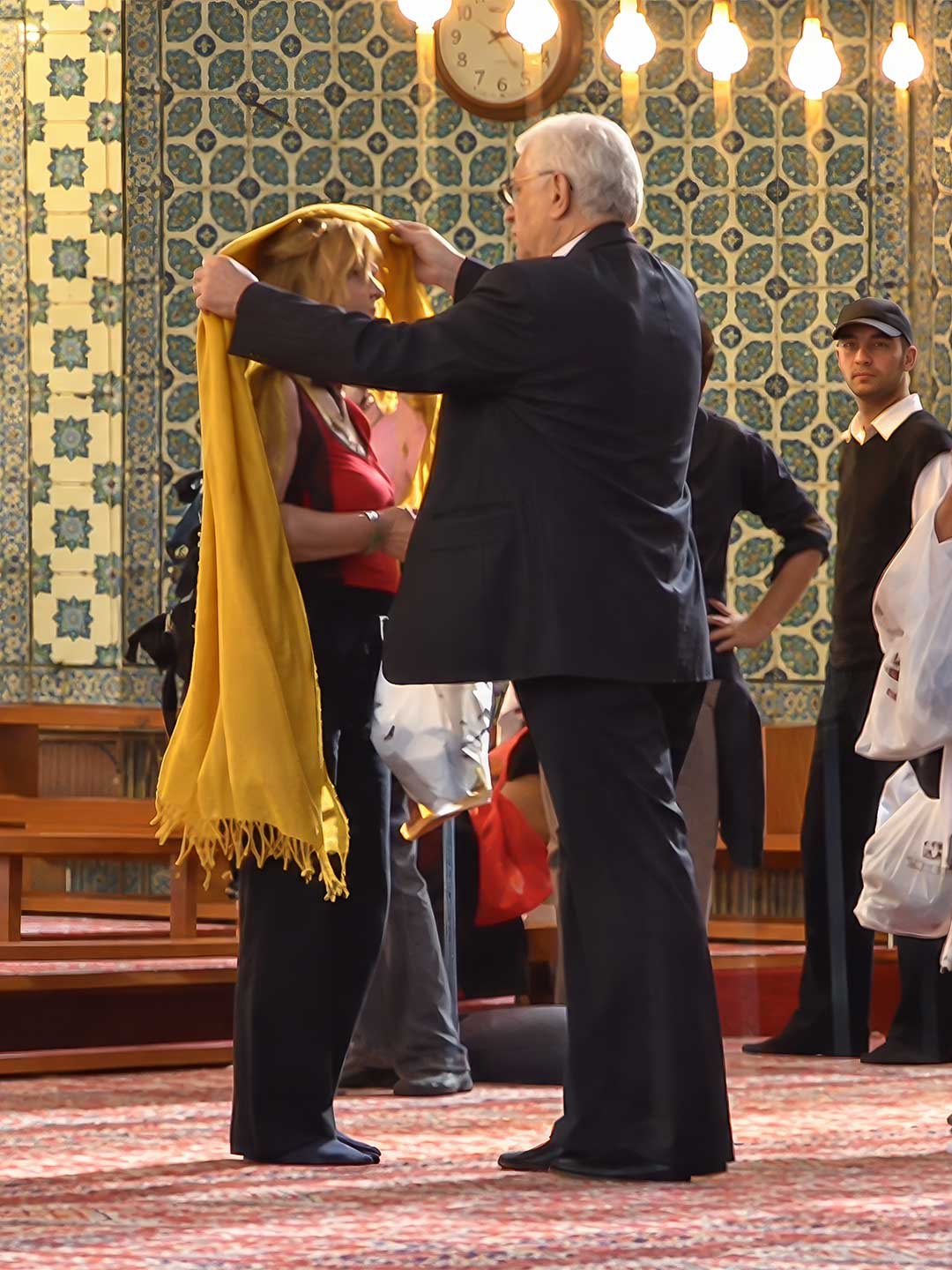 Istanbul-Blaue-Moschee-Bekleidung Ein Mann im Anzug legt einer Frau in einer Moschee ein gelbes Kopftuch um.