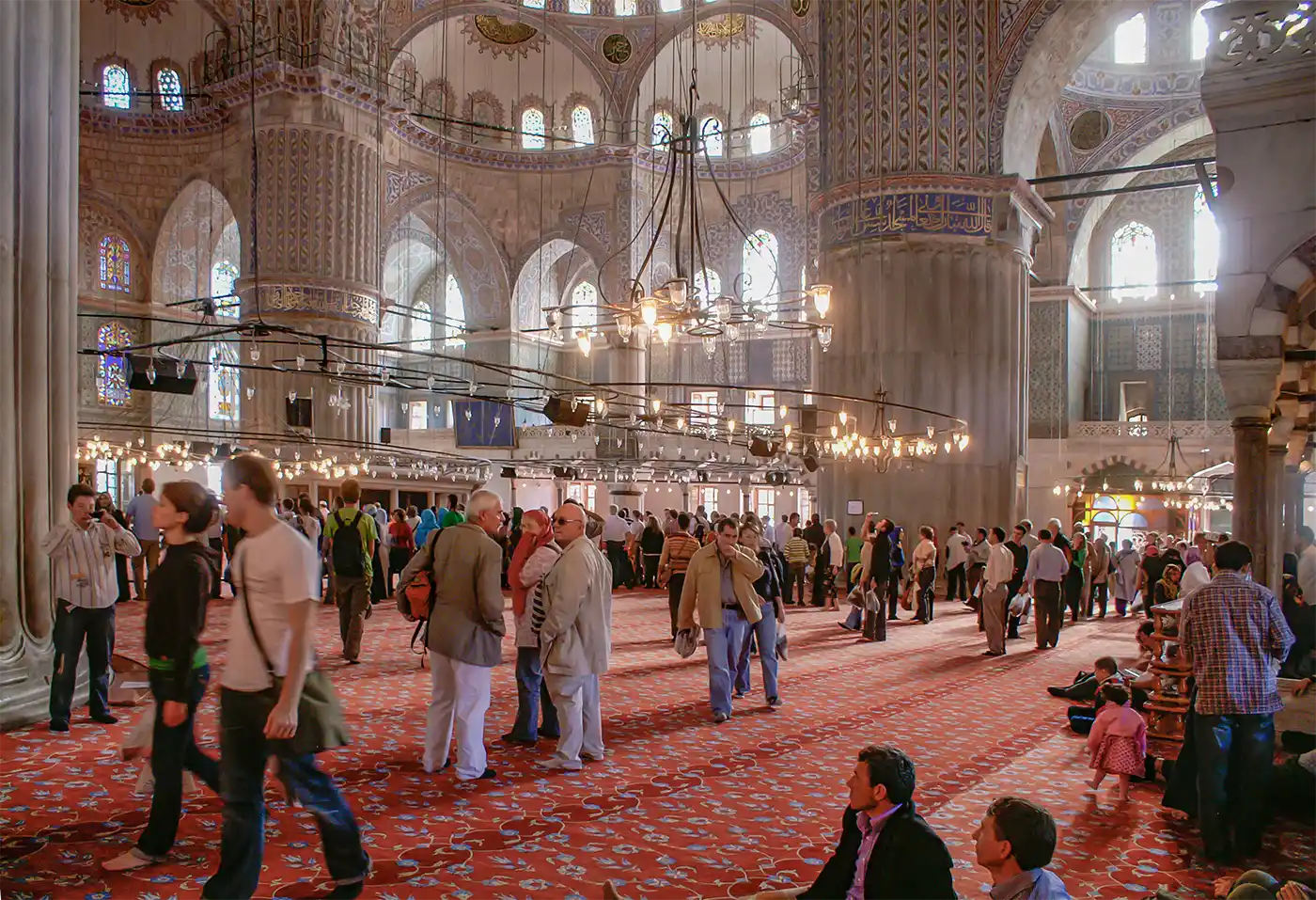 Istanbul-Blaue-Moschee Innenansicht der Blauen Moschee in Istanbul, in der Touristen auf einem großen, rot gemusterten Teppich spazieren gehen.