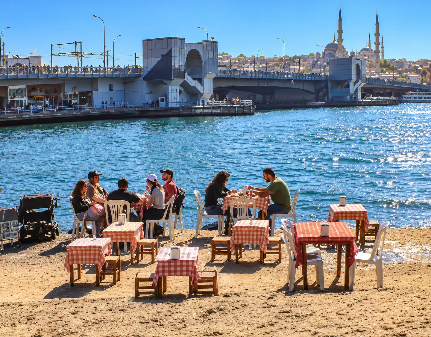 Istanbul-Galata-Bruecke-Beach-Bar Menschen, die an Tischen an einem sandigen Ufer sitzen, im Hintergrund eine Brücke und eine Moschee.