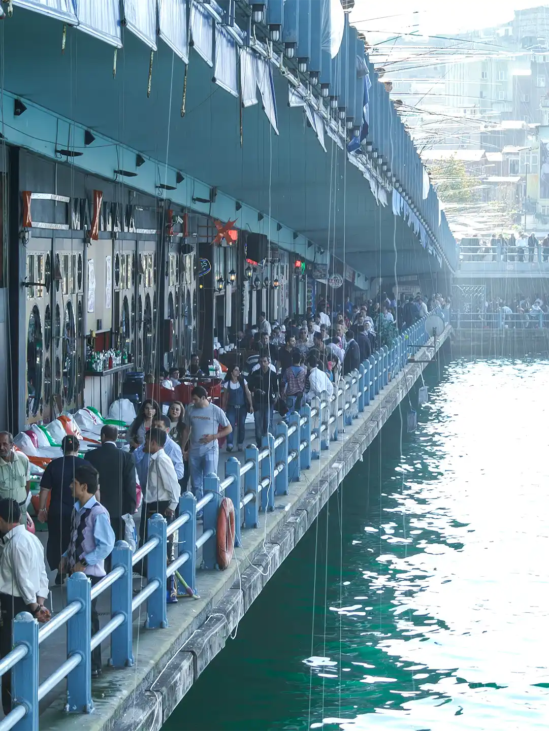 Istanbul-Galata-Bruecke-Plattform Menschen, die unter einer Brücke entlang einer belebten Uferpromenade spazieren gehen.