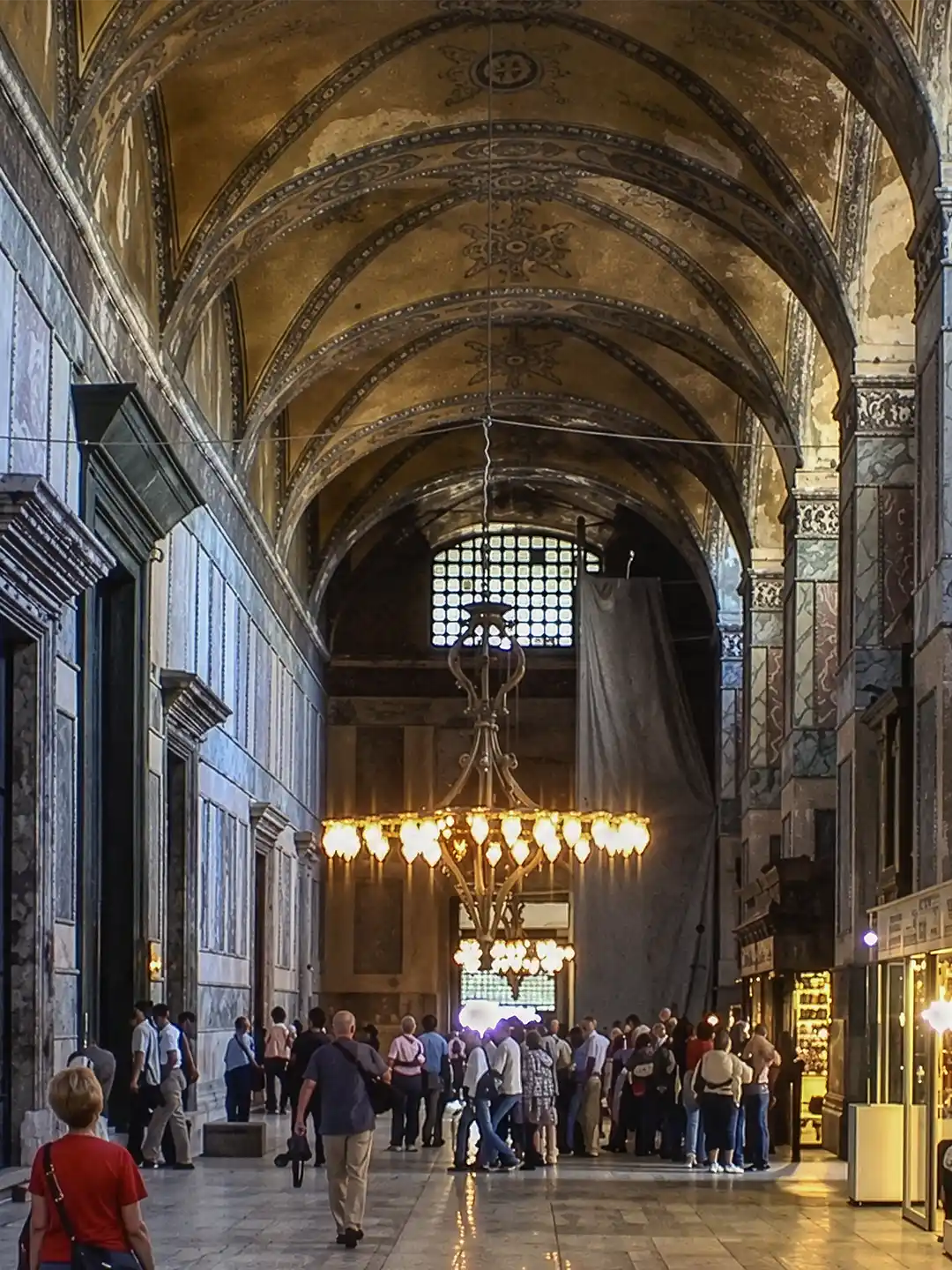 Istanbul-Hagia-Sophia-Seitengewoelbe Das Innere der Hagia Sophia mit hohen Gewölbedecken, einem großen Kronleuchter und Besuchern, die durch den Saal schlendern.
