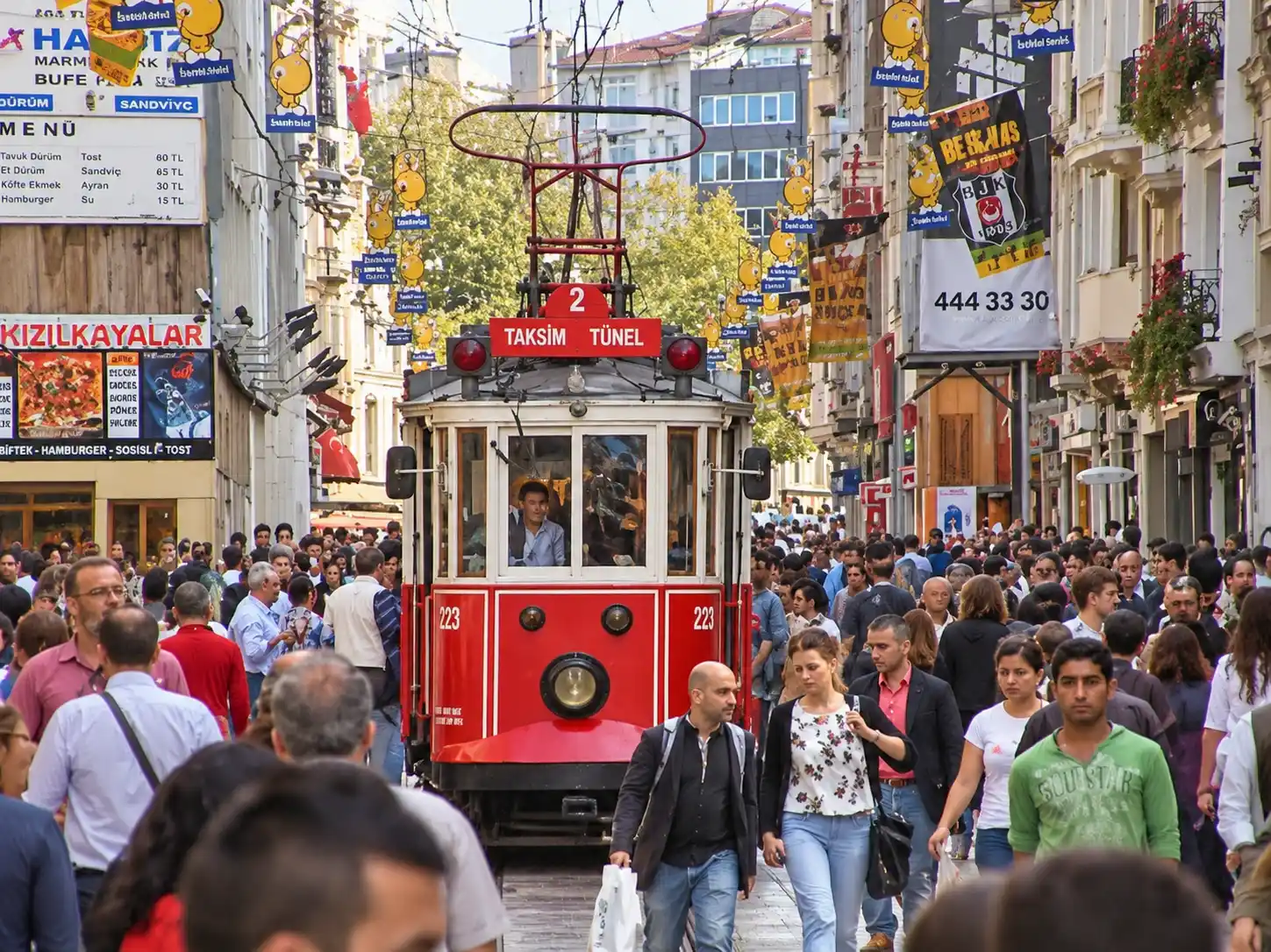 Istanbul-Strassenbahn Eine historische rote Straßenbahn fährt durch eine belebte Fußgängerzone in Istanbul, die von Geschäften und Werbebannern gesäumt ist.