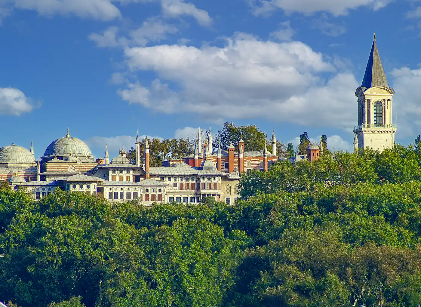 Istanbul-Topkapi-Palast Der Topkapi-Palast mit seinen Kuppeln und Türmen, die sich unter blauem Himmel über dichten, grünen Bäumen erheben.