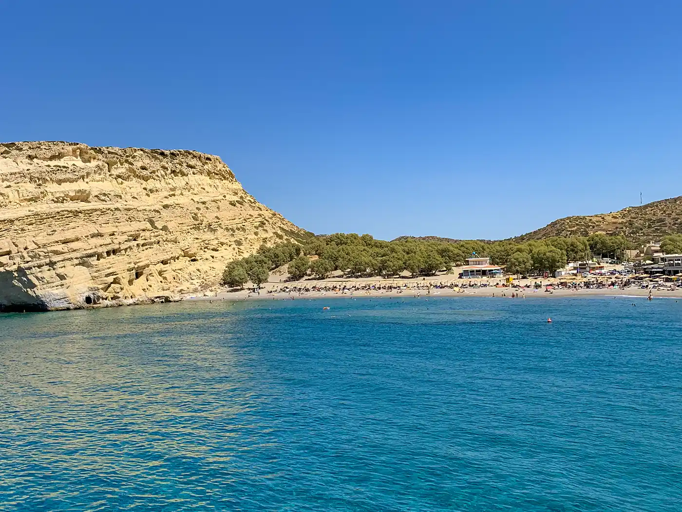 Matala-Beach-Hoehlen Links die Höhlen von Matala in beigefarbenen Klippen, daneben der Sandstrand mit Badenden am Meer unter einem strahlend blauen Himmel.