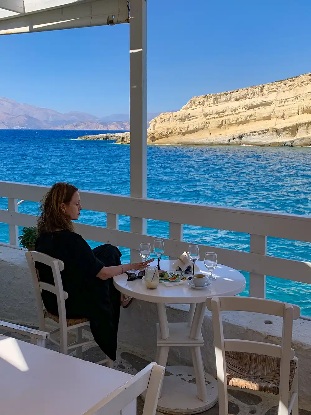 Eine Frau sitzt an einem Tisch auf einem Balkon mit Blick auf das strahlend blaue Meer und die felsigen Klippen.