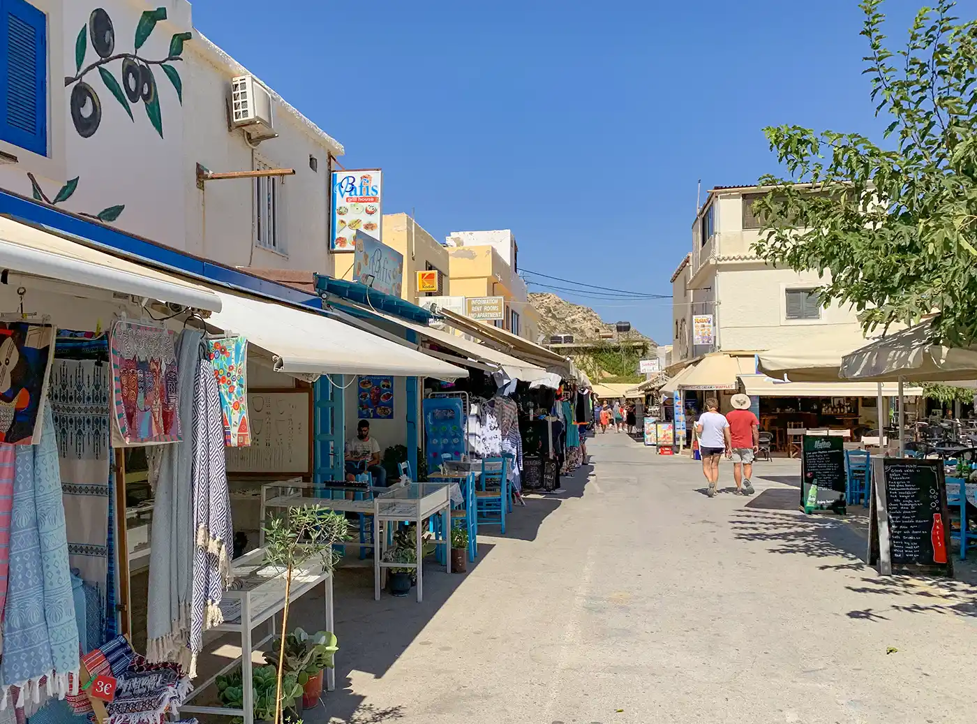Fußgängerzone in einem griechischen Dorf, gesäumt von Geschäften, Cafés und aufgehängten Textilien.