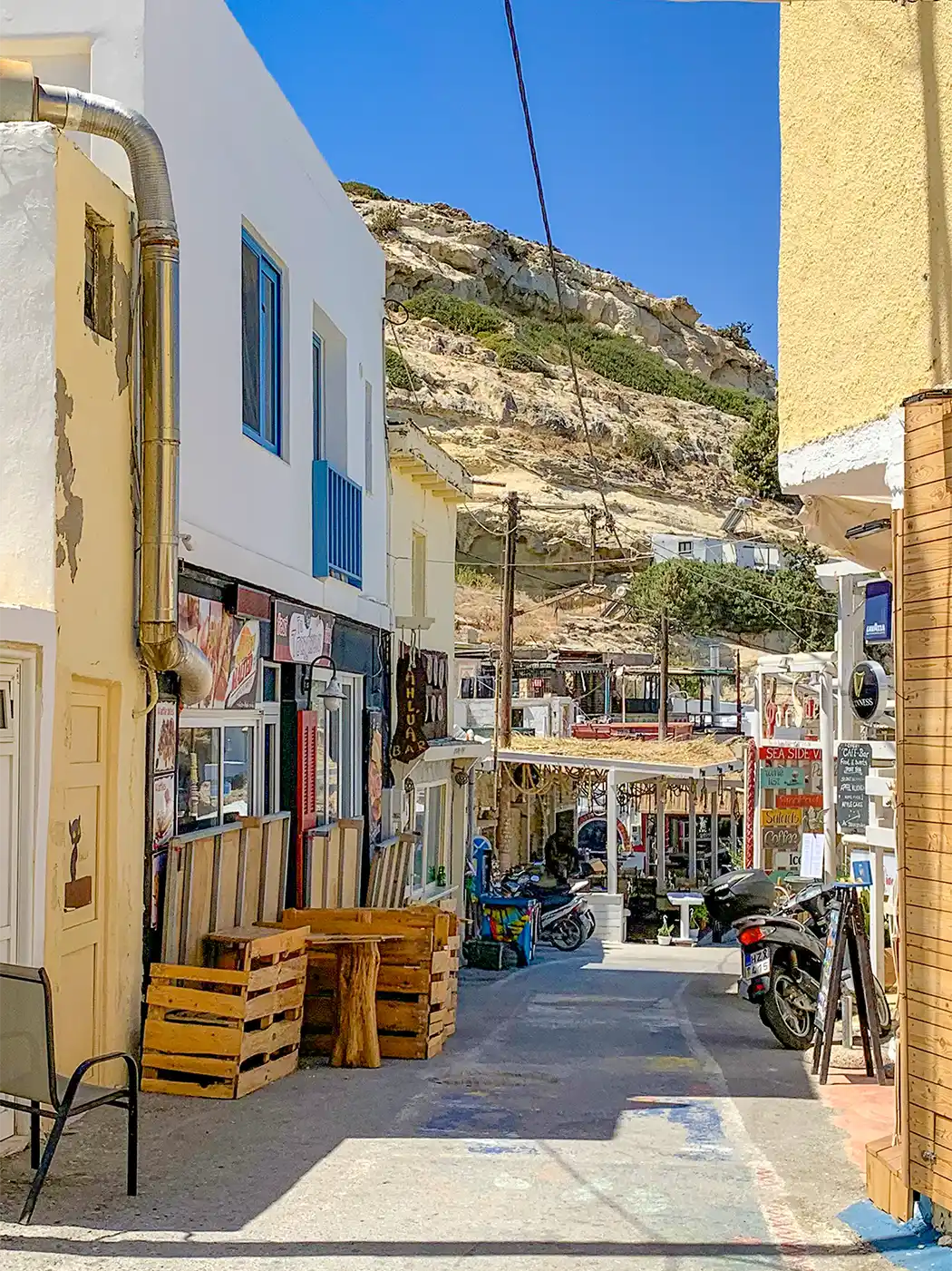 Matala-Souvenirshops Eine schmale Straße in einer Küstenstadt, gesäumt von kleinen Geschäften, Cafés und Terrassenmöbeln aus Holz.