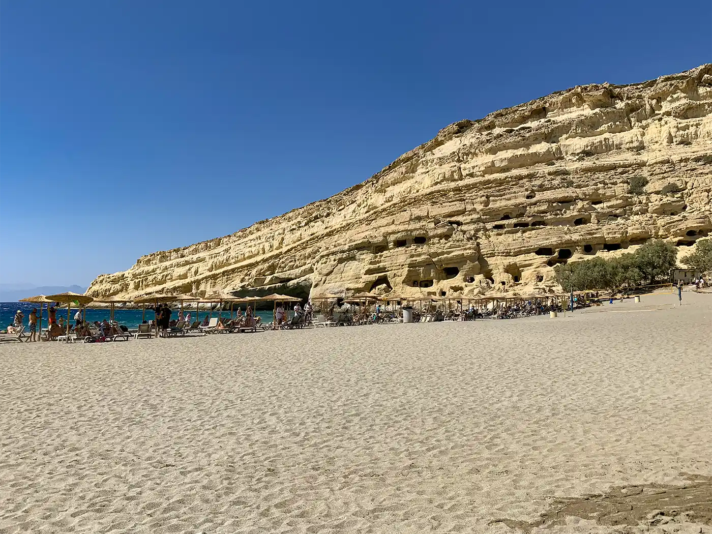 Menschen, die sich auf Strandliegen unter Sonnenschirmen am Fuße einer großen Klippe mit ausgehöhlten Höhlen entspannen.