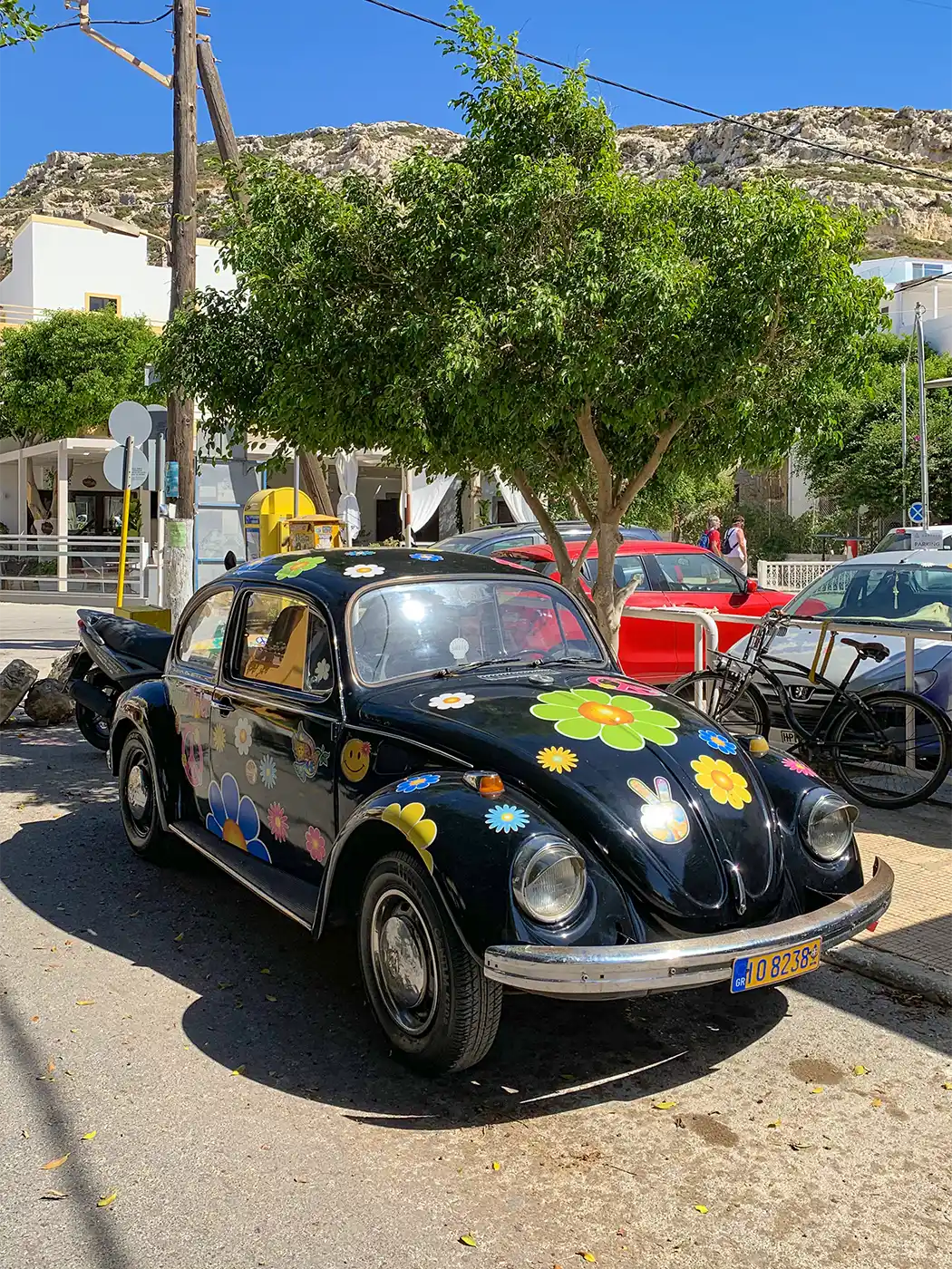 Matala-VW-Kaefer Ein schwarzer Volkswagen Käfer, verziert mit bunten Blumen- und Hasenaufklebern, steht unter einem Baum in Matala.