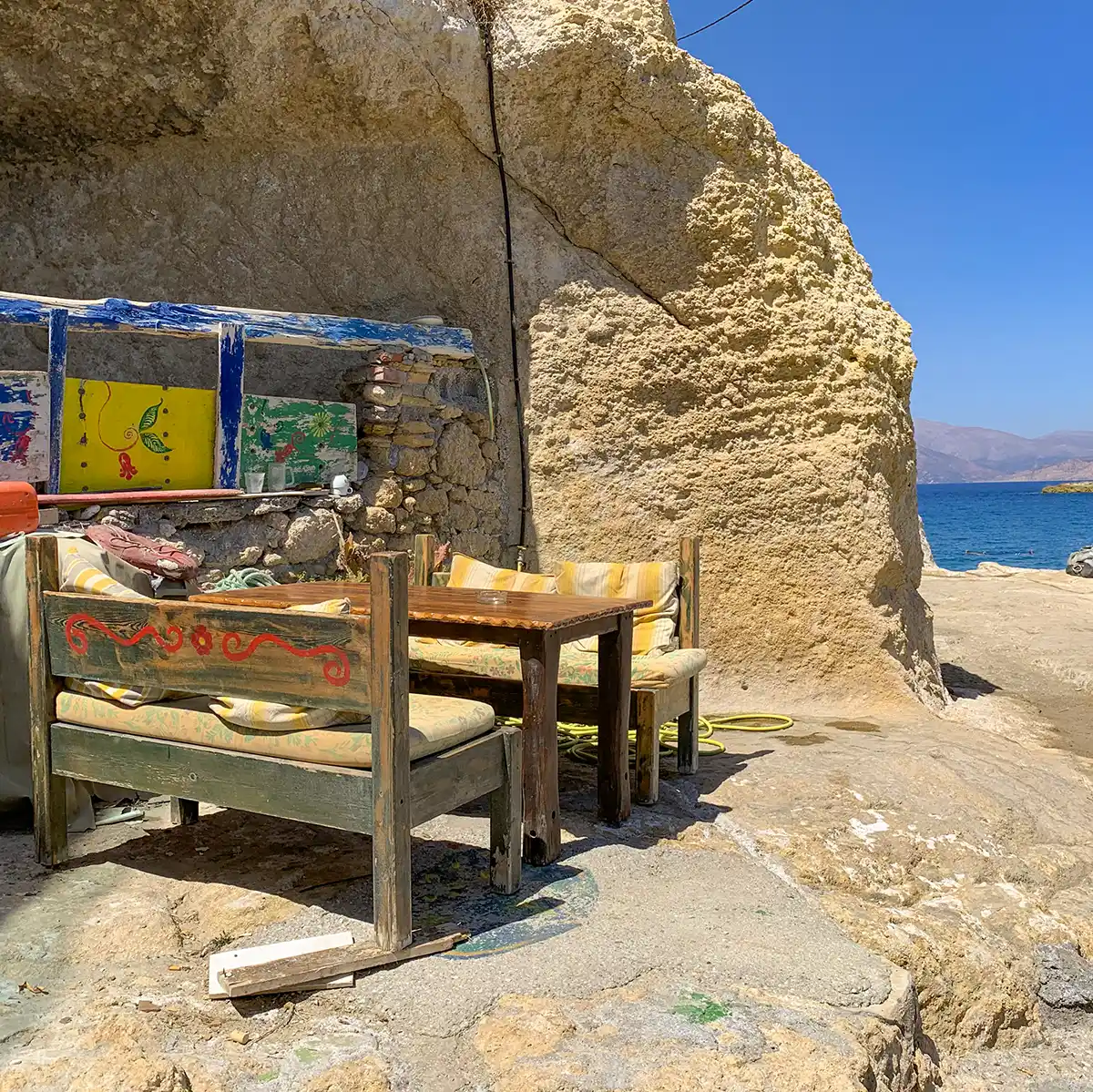 Ein Holztisch und Bänke mit Kissen, die unter einem Felsvorsprung in Matala am Meer stehen.