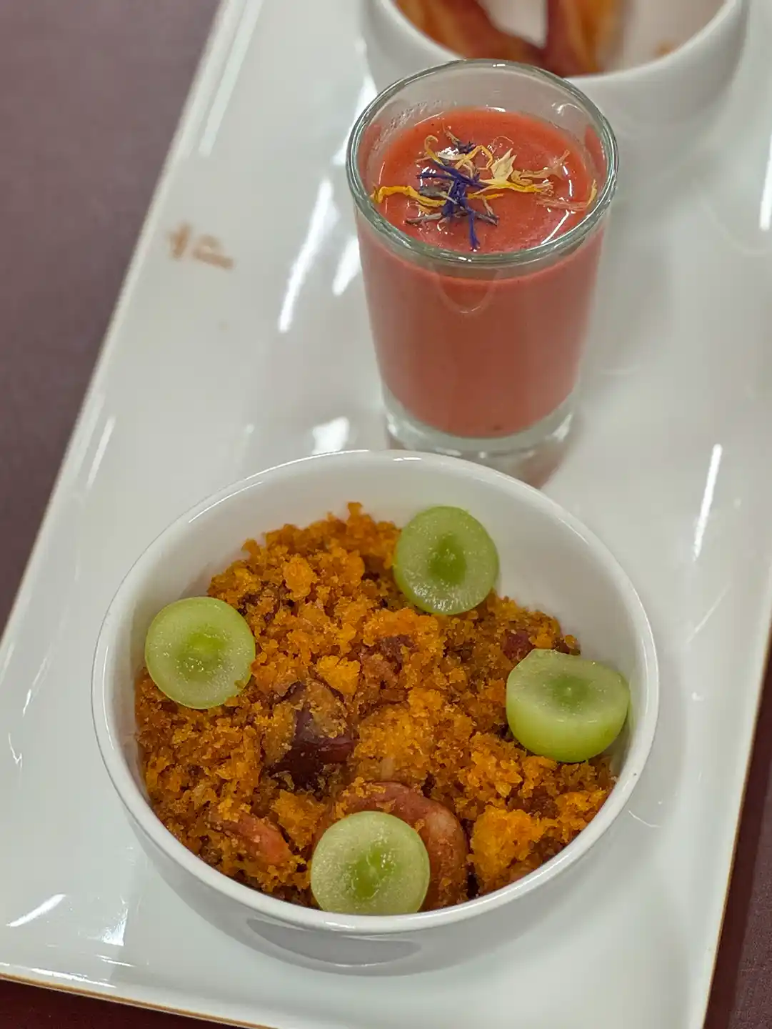 Migas-Spezialitaeten-Kastilien-La-Mancha Eine Schüssel Migas, garniert mit grünen Traubenscheiben, daneben ein kleines Glas Gazpacho, verziert mit Blütenblättern.