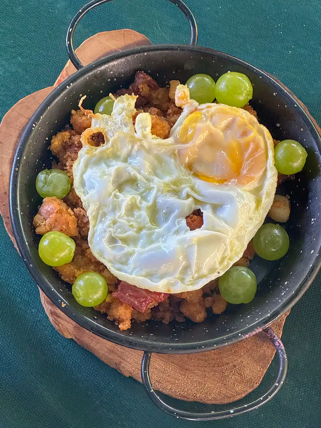 Migas-Spiegelei-Spezialitaeten-Kastilien-La-Mancha Ein Spiegelei, serviert auf Migas mit grünen Trauben in einer kleinen Metallpfanne.